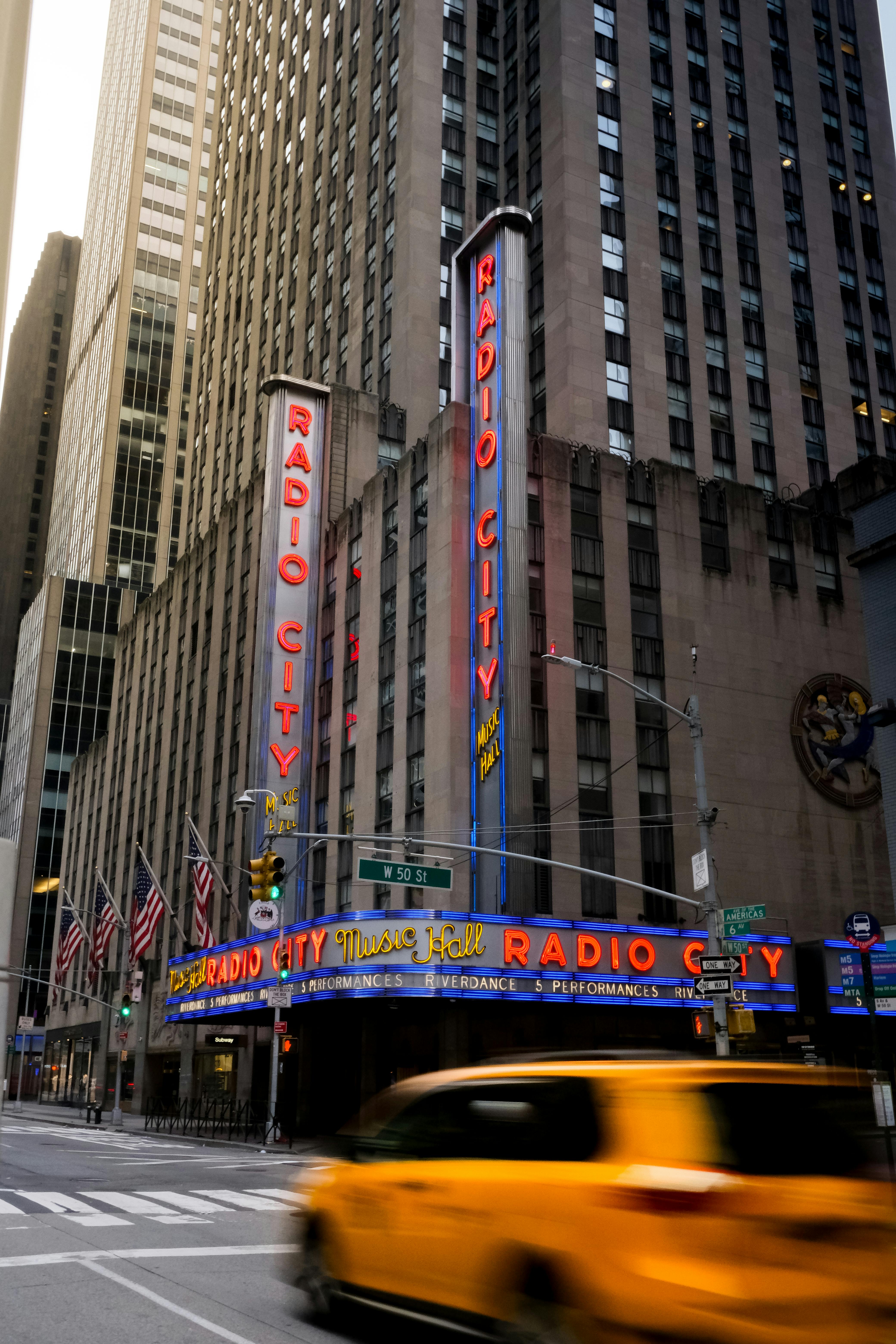 Kostenlos Farbenfrohe Ansicht der Radio City Music Hall mit einem fahrenden gelben Taxi, die die Essenz von New York City einfängt. Stock-Foto