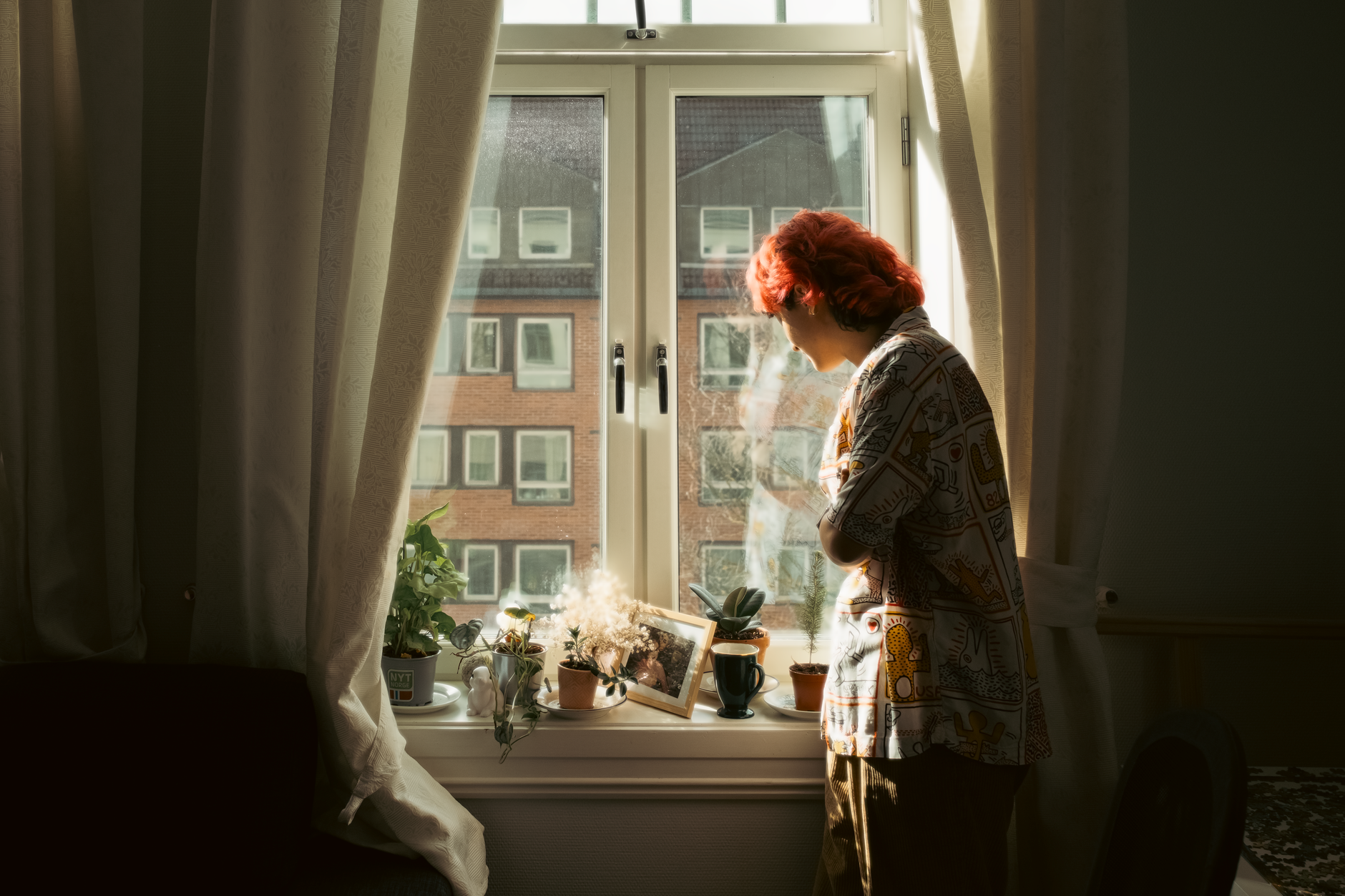 A woman stands thoughtfully by a sunlit window, glancing to the side