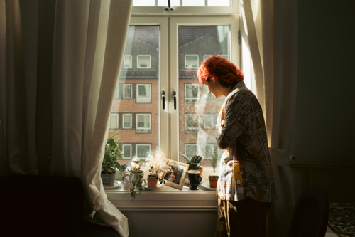 Free A woman stands thoughtfully by a sunlit window with plants. Stock Photo
