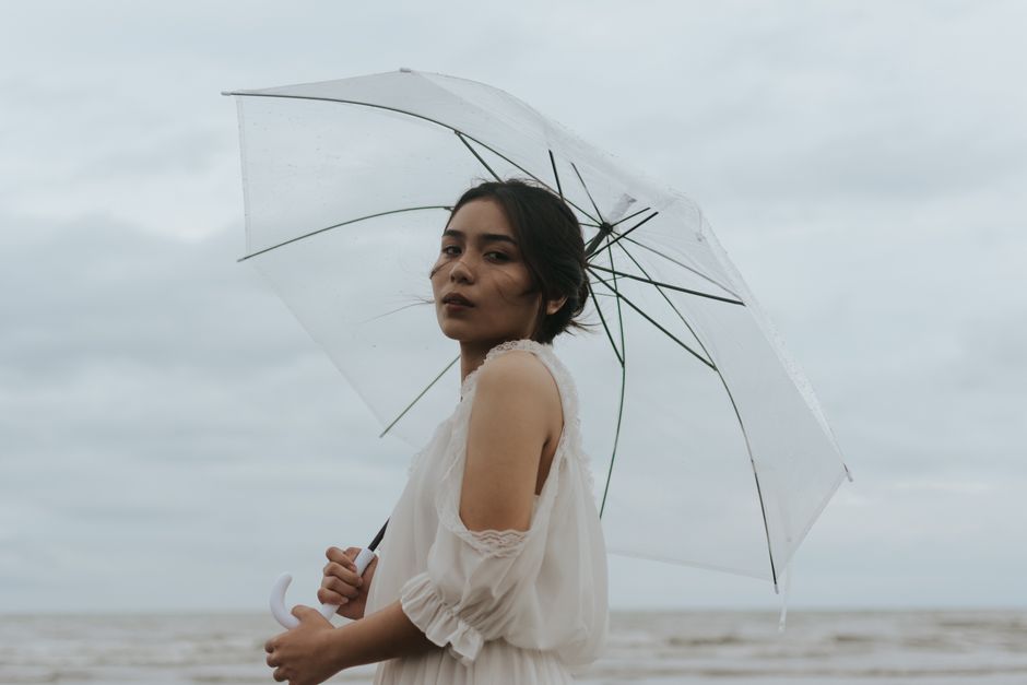 A stylish woman poses with a transparent umbrella by the sea on a cloudy day, exuding elegance and charm.