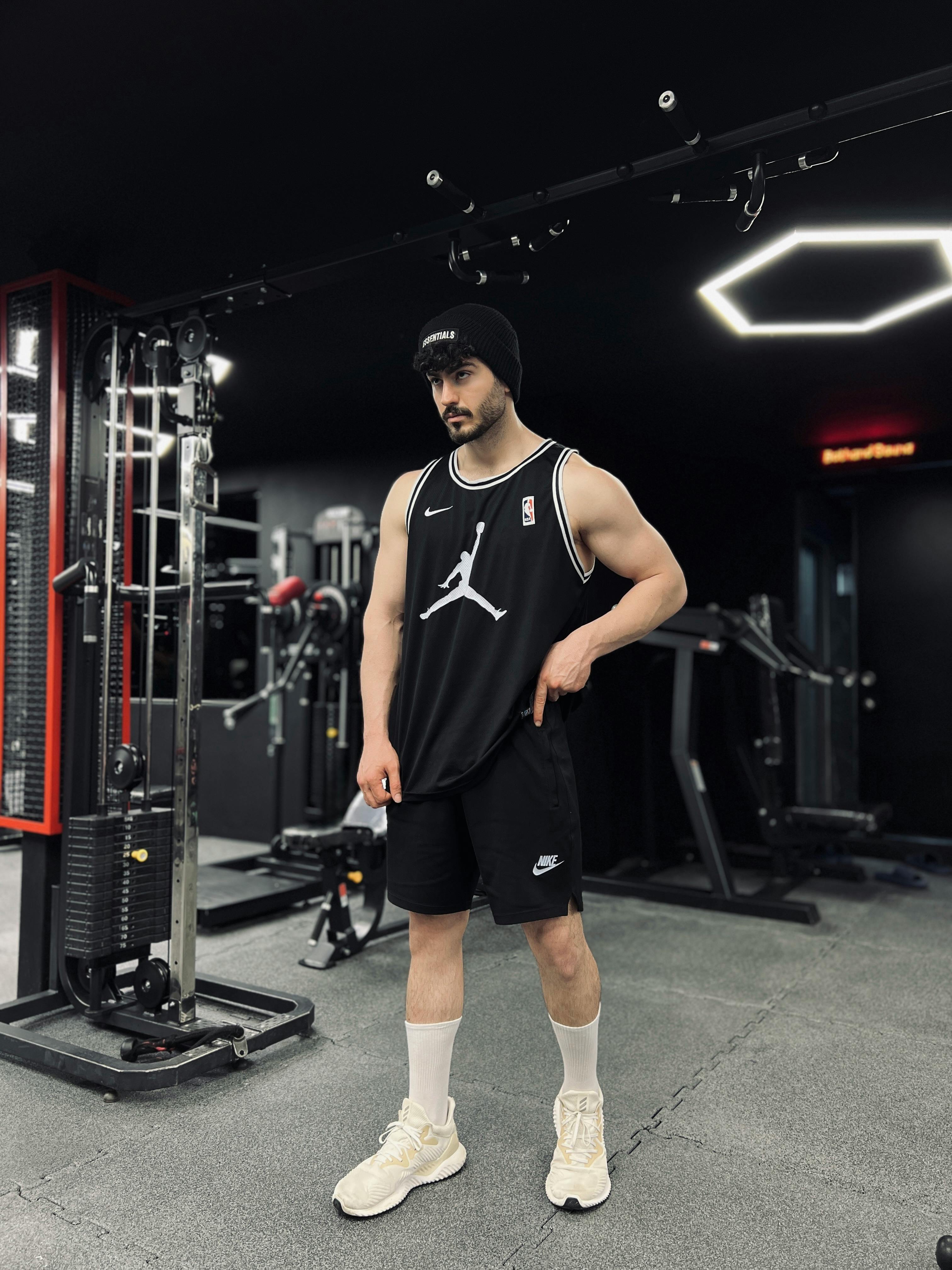 A man in a gym, wearing black sports attire, posing confidently with exercise equipment in background.