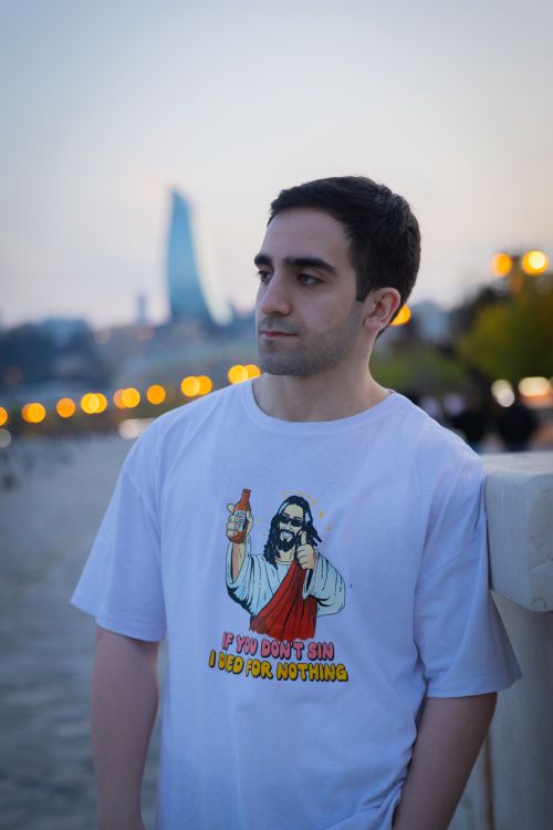 Free Young man wearing graphic t-shirt stands by waterfront at dusk, city lights in background. Stock Photo