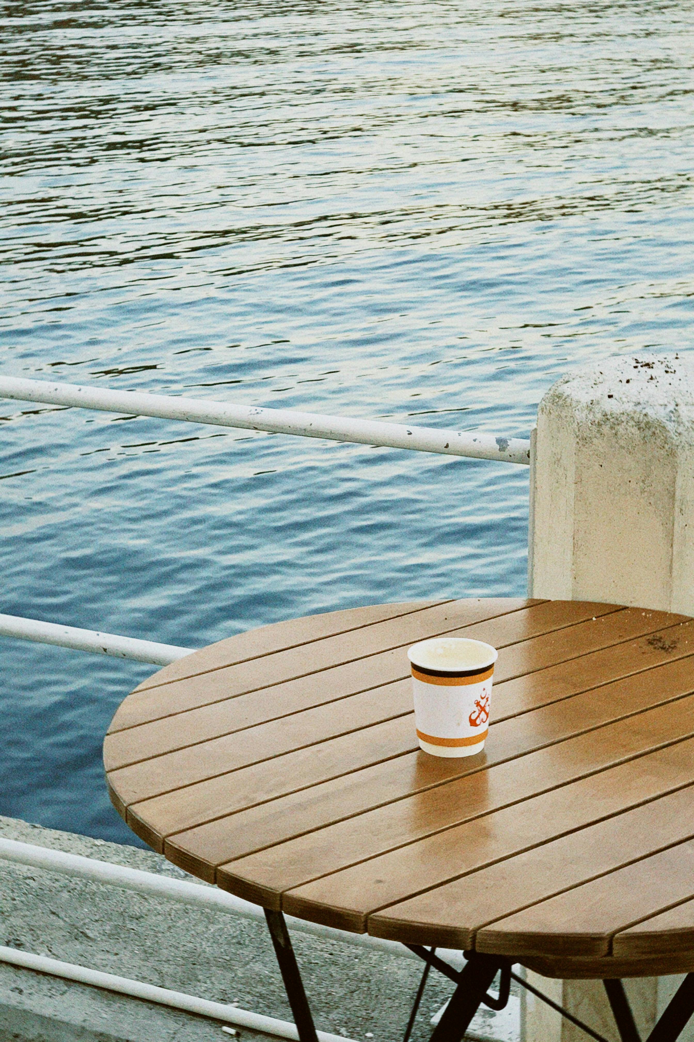A lone coffee cup sits on a wooden table by the calm waterfront.
