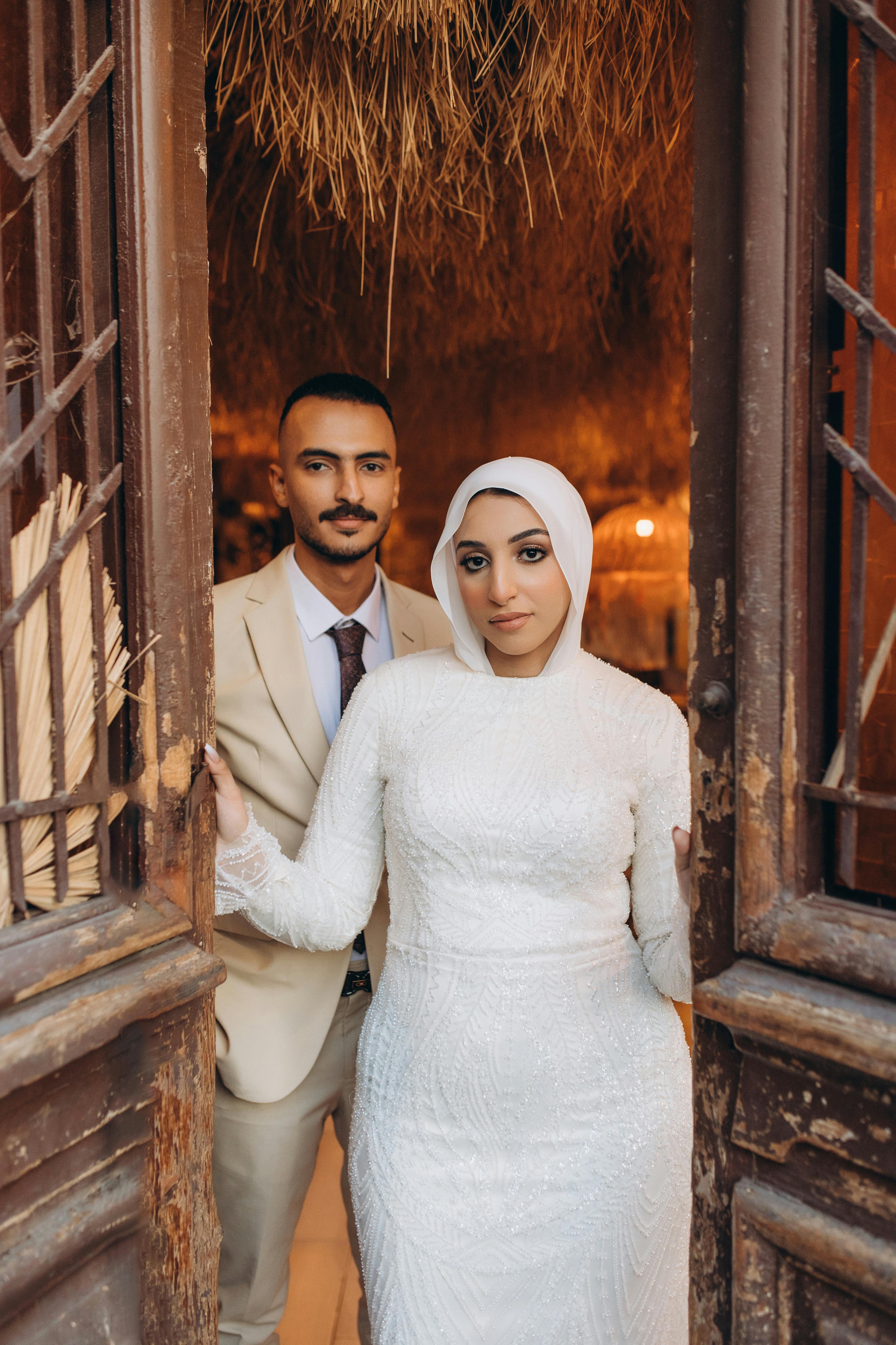Charming couple in traditional wedding attire standing at a rustic wooden doorway.