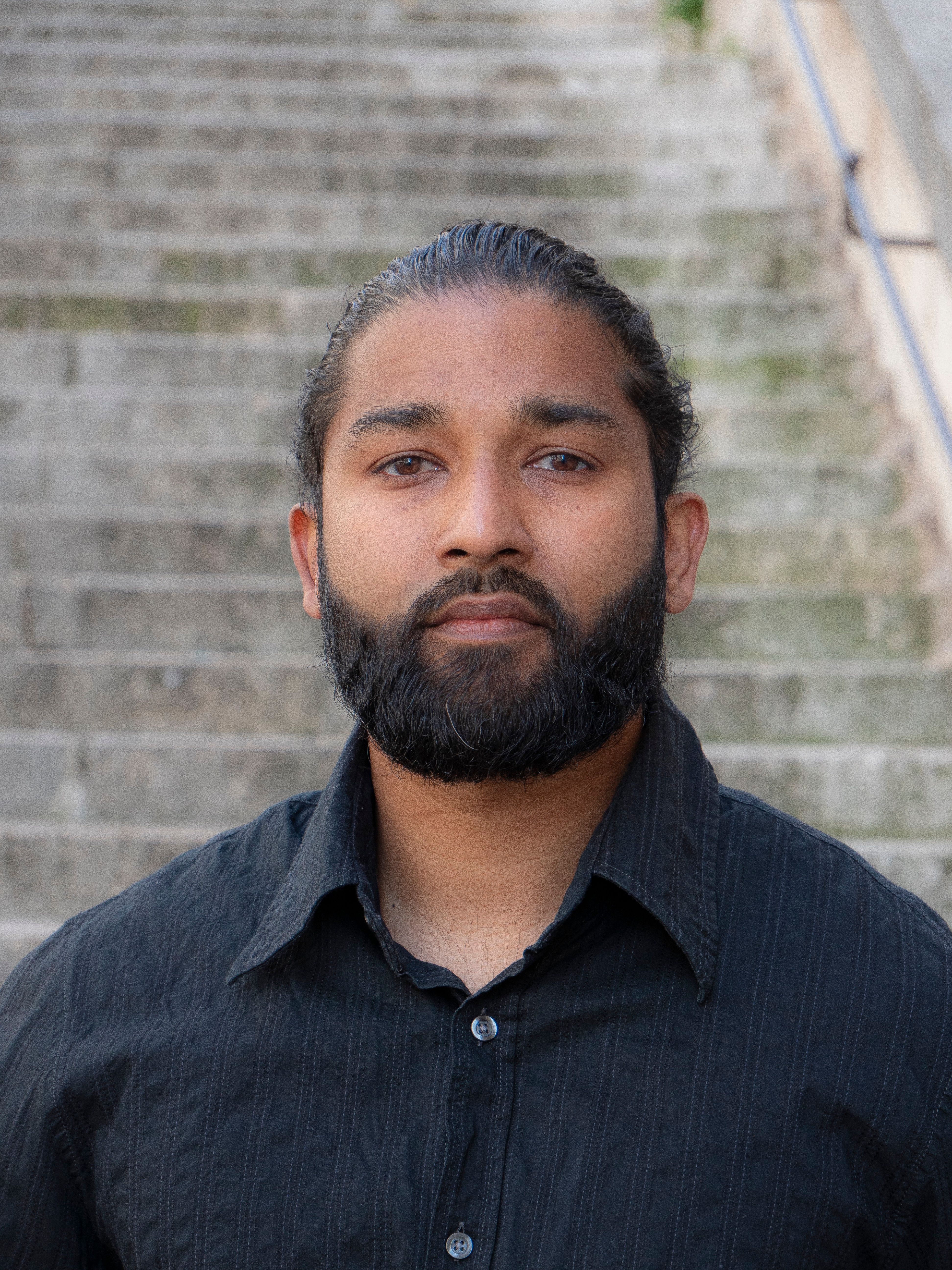 Bearded adult man in black shirt standing on outdoor stairs, looking serious.