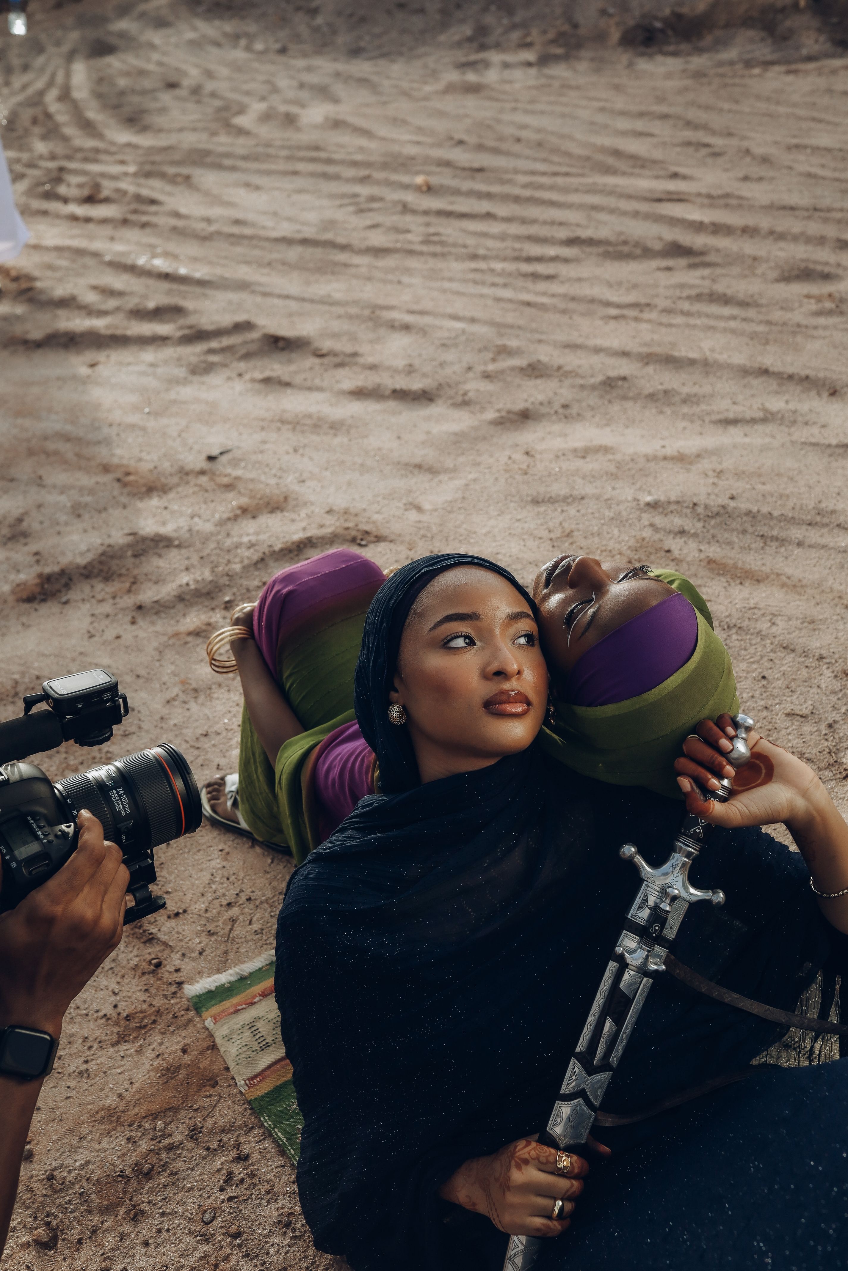A woman in traditional attire poses with a sword on sandy terrain, watched by a cameraman.