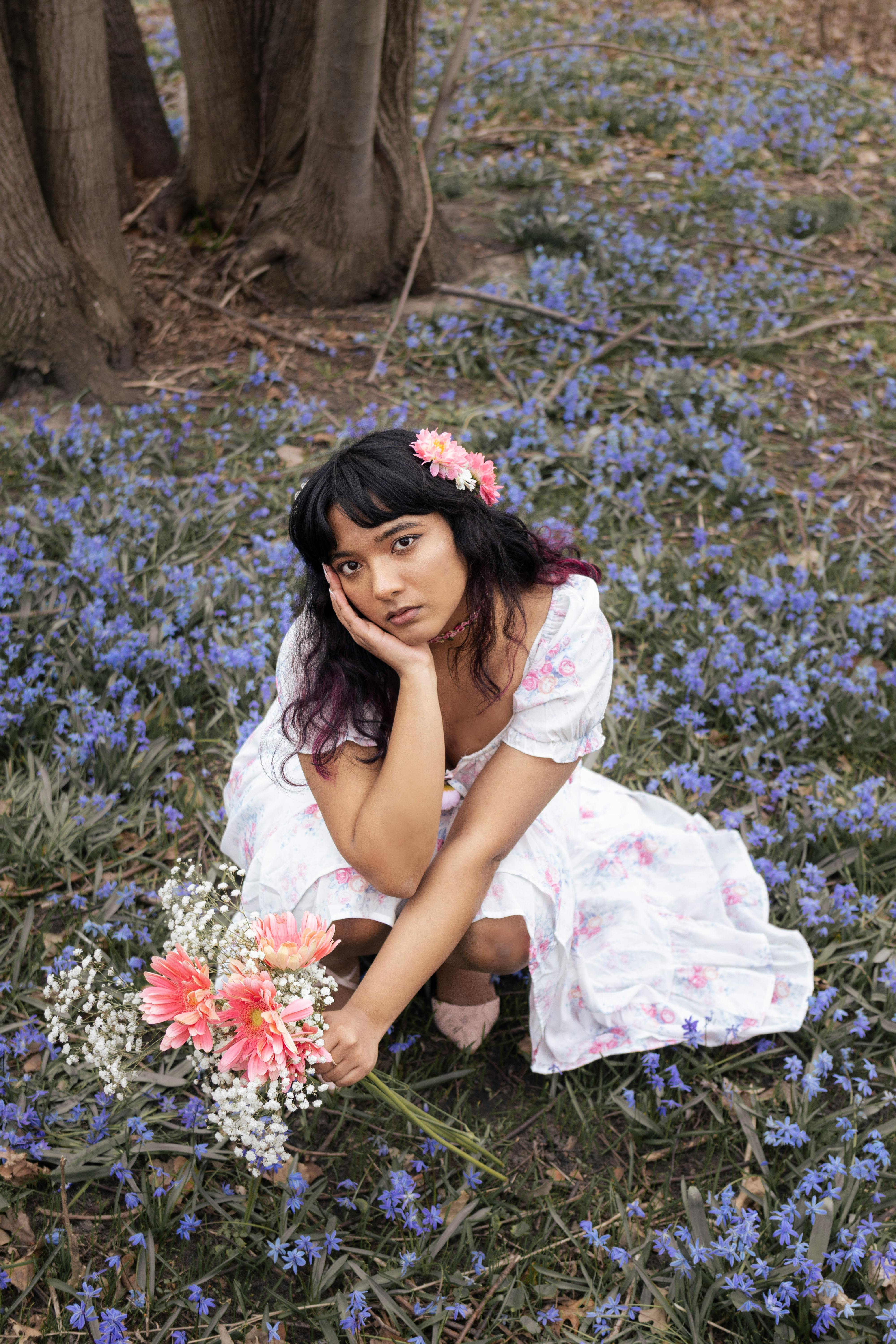 Woman in a white floral dress with pink flowers kneeling among blue flowers outdoors.