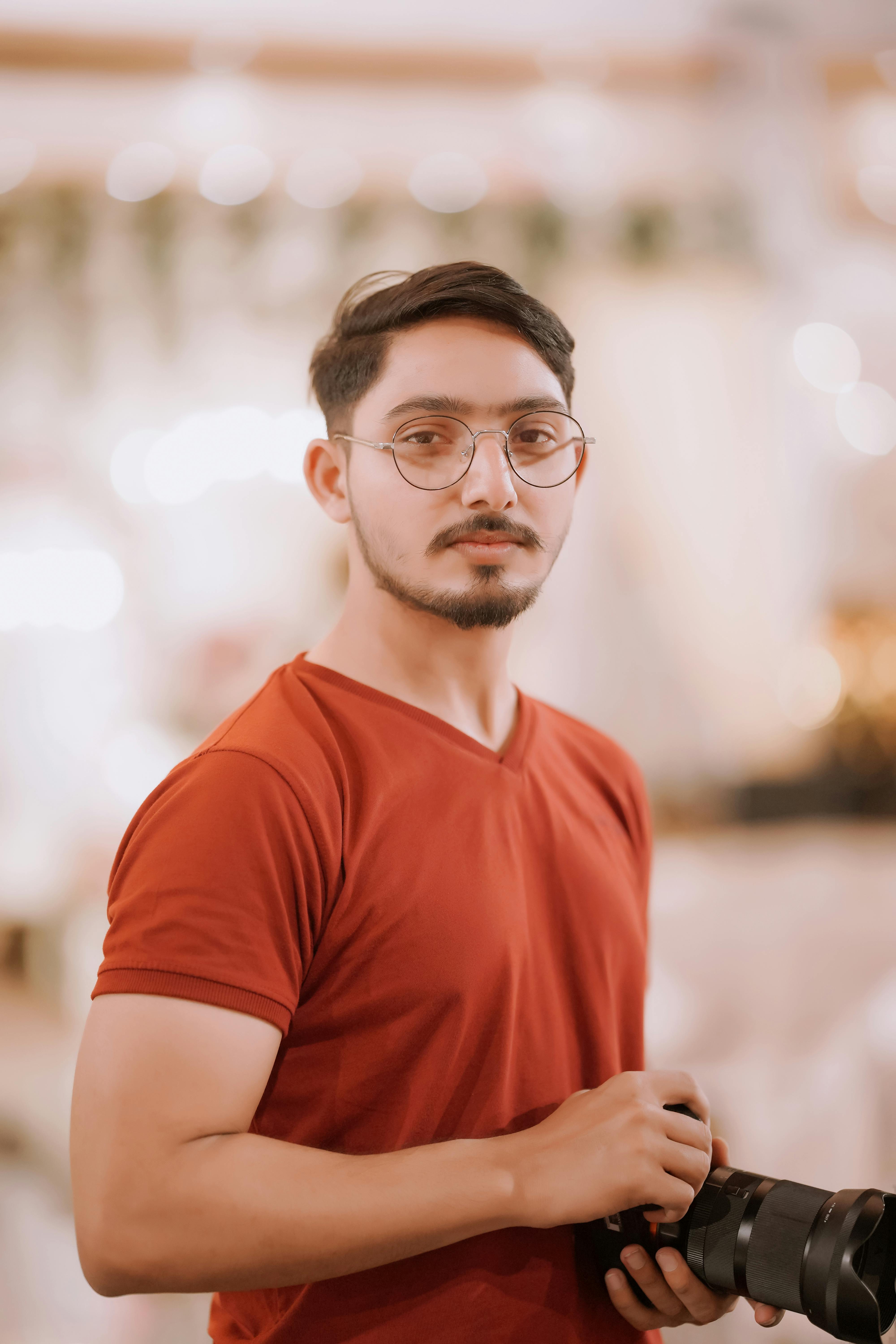 Miễn phí Một nhiếp ảnh gia nam trẻ tuổi đang cầm máy ảnh, đứng trong nhà với phông nền mờ. Ảnh lưu trữ