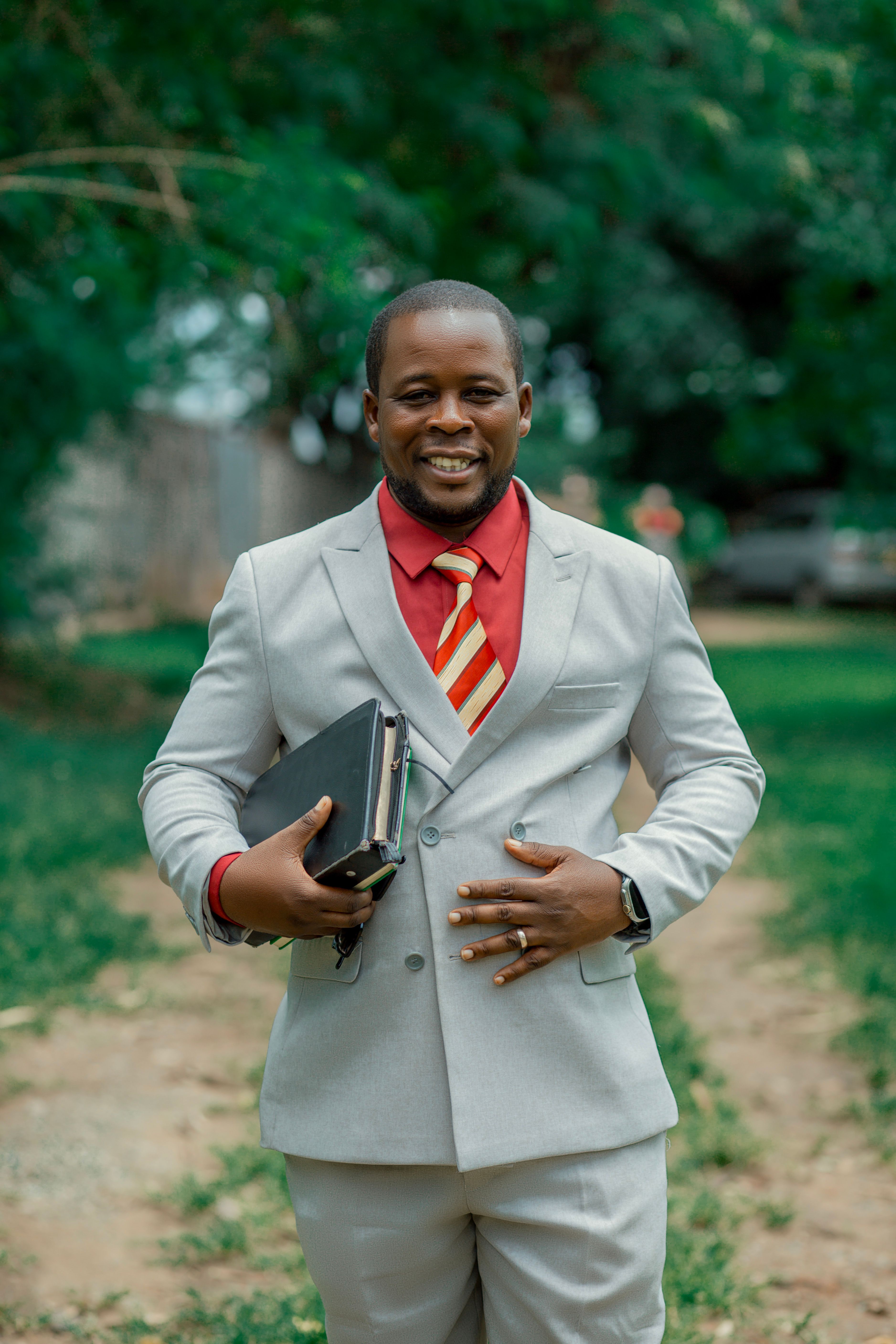 Smiling man in a grey suit with a red shirt holding a folder outdoors.