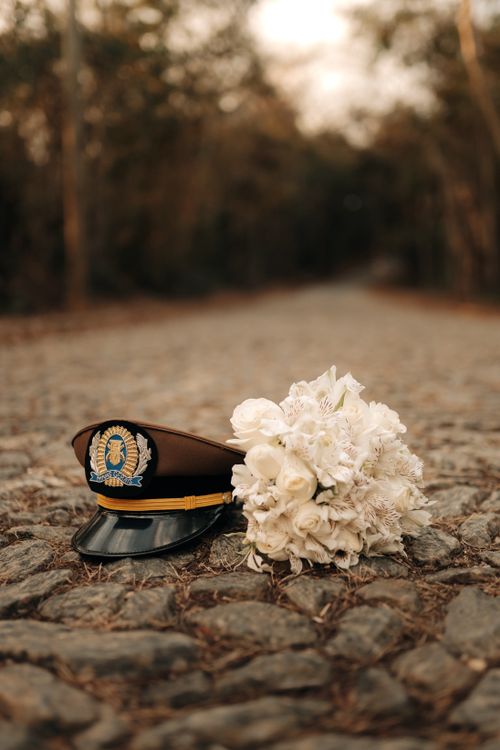 Free A military cap and white floral bouquet sit on a cobblestone path in a serene outdoor setting. Stock Photo
