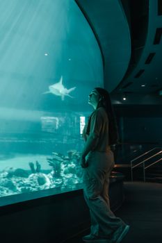 A woman gazing at a shark swimming gracefully in an indoor aquarium exhibit, creating a serene and reflective moment.