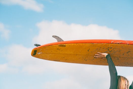 A surfer lifts a colorful board under a clear blue sky, embracing the adventurous lifestyle.