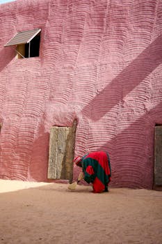 A person in traditional attire sweeps in front of a textured pink clay wall under bright sunlight.
