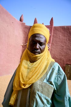 Portrait of man wearing vibrant traditional attire against adobe wall in desert landscape.