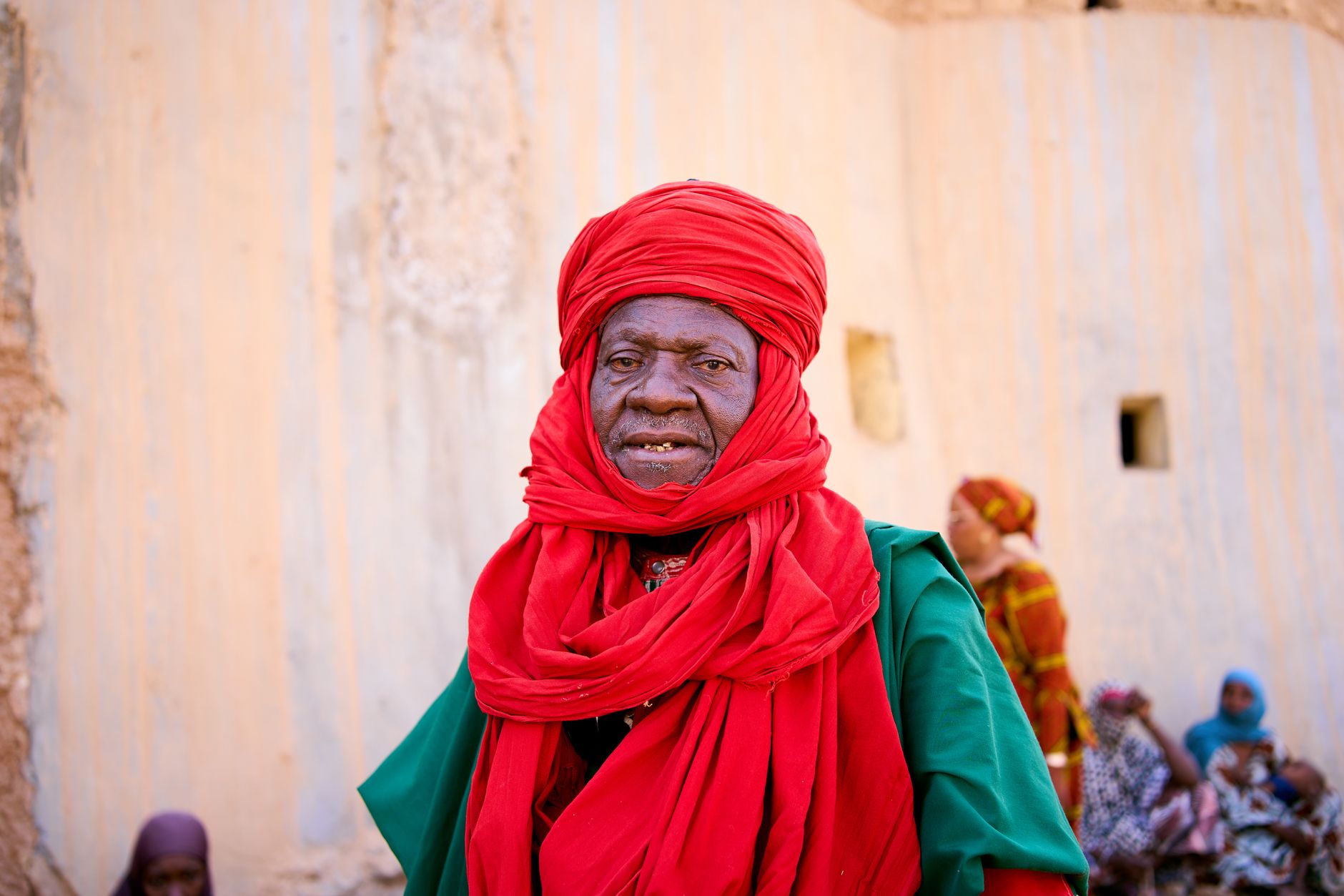 An elderly man in traditional red attire stands confidently outdoors against a textured wall.