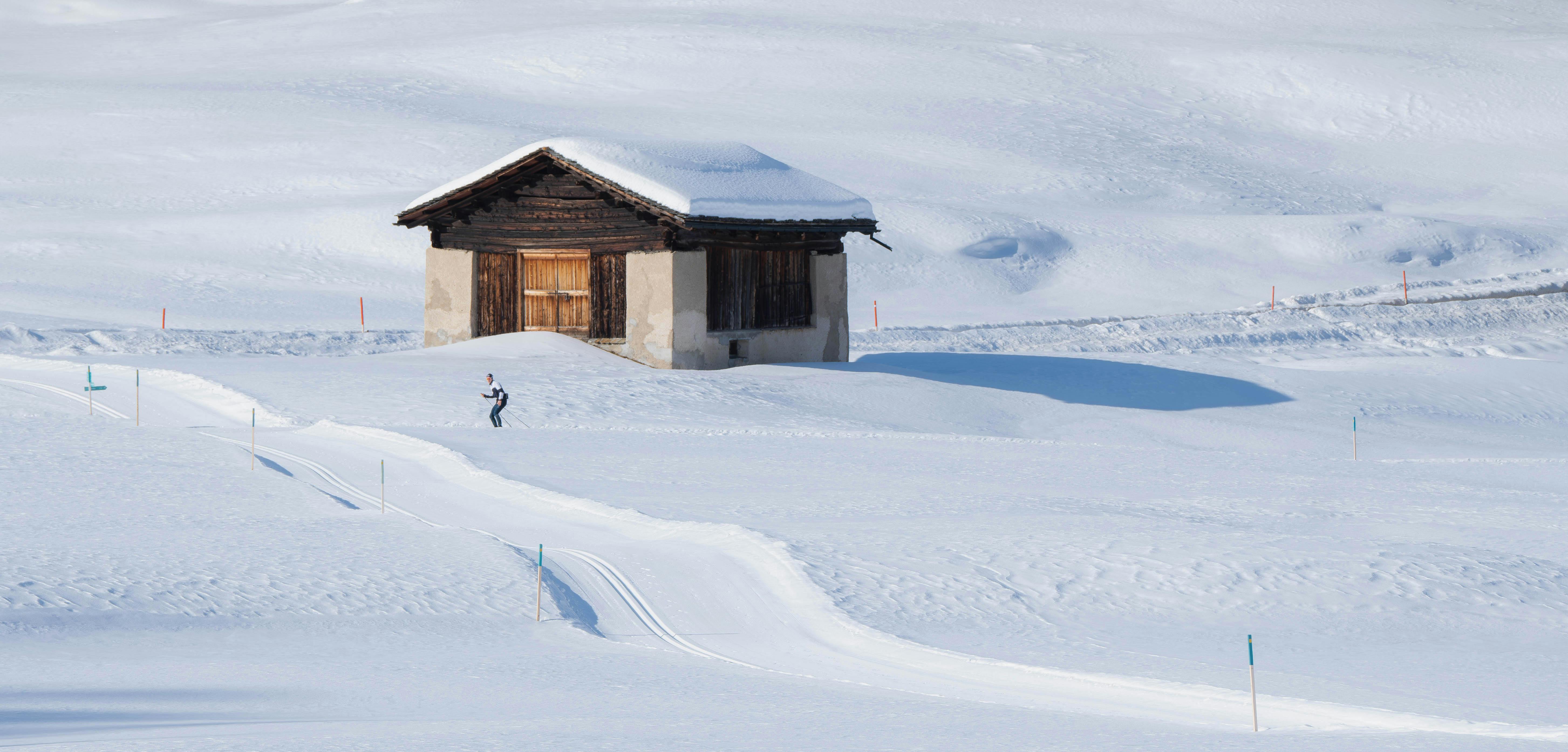 A lone skier glides past a rustic cabin in a vast, snowy landscape, capturing winter's serenity.