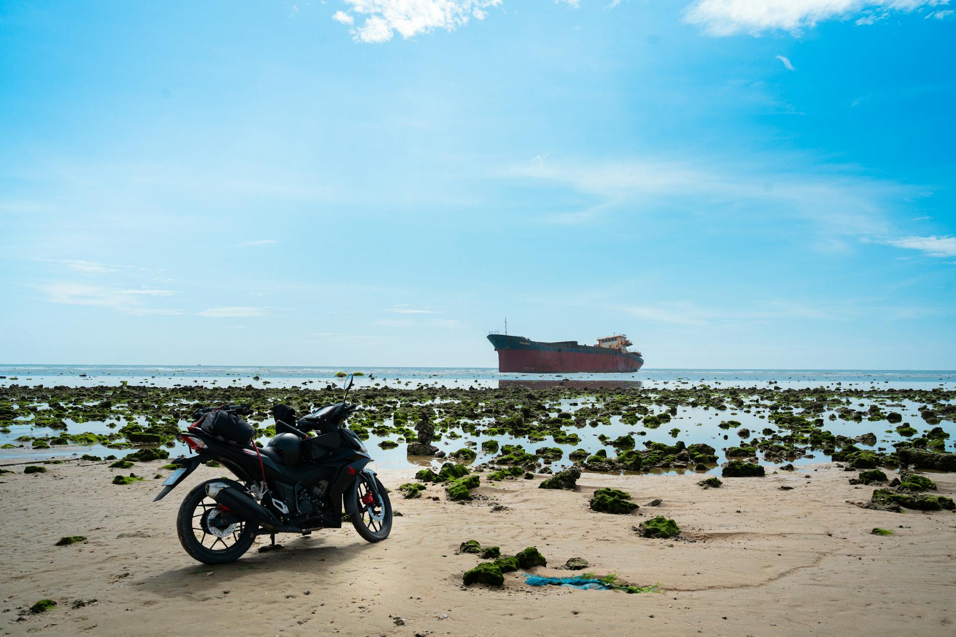 A motorcycle parked on a sandy beach with a distant cargo ship and clear skies.