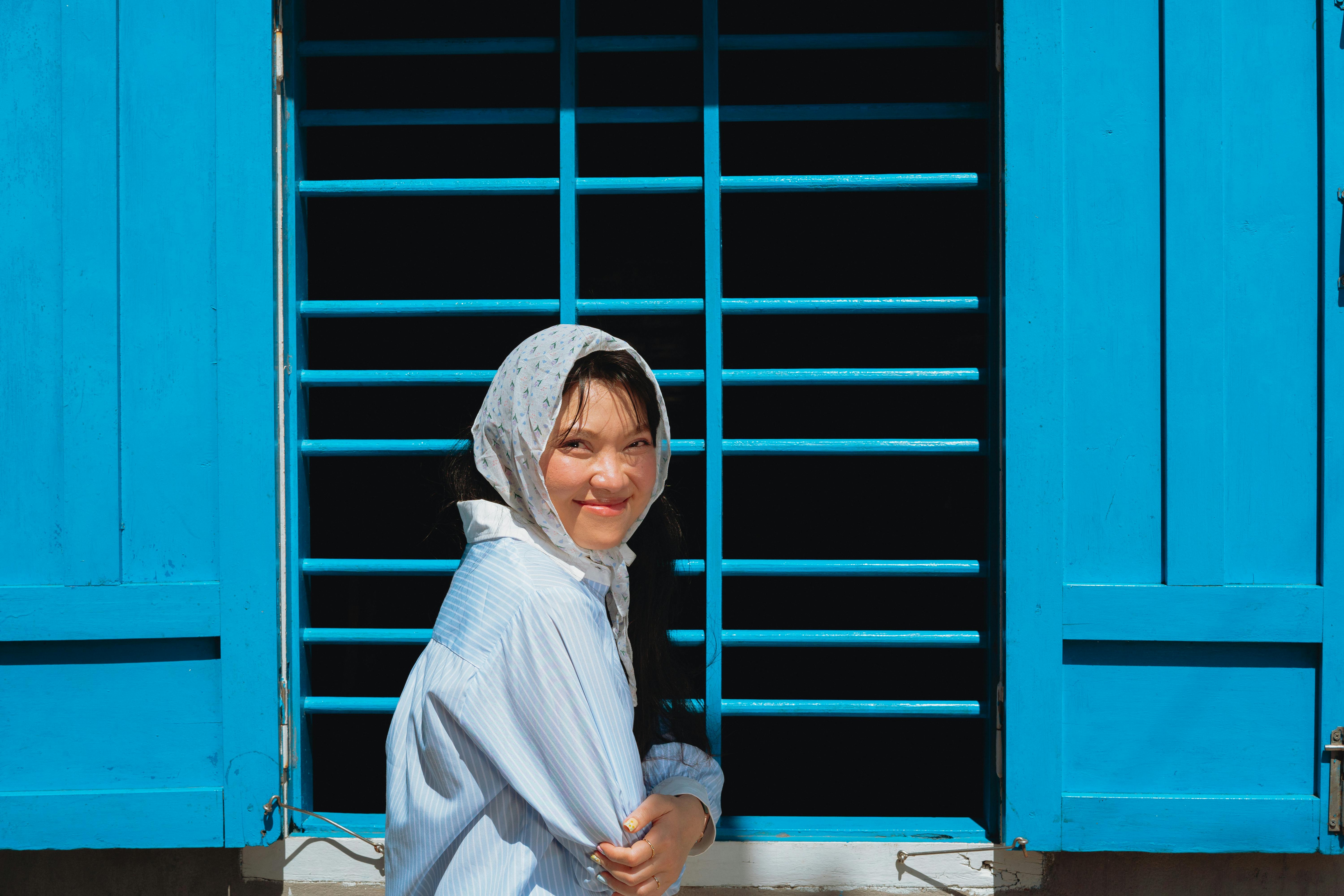 A cheerful woman in a headscarf stands by a vibrant blue window, exuding warmth and charm.