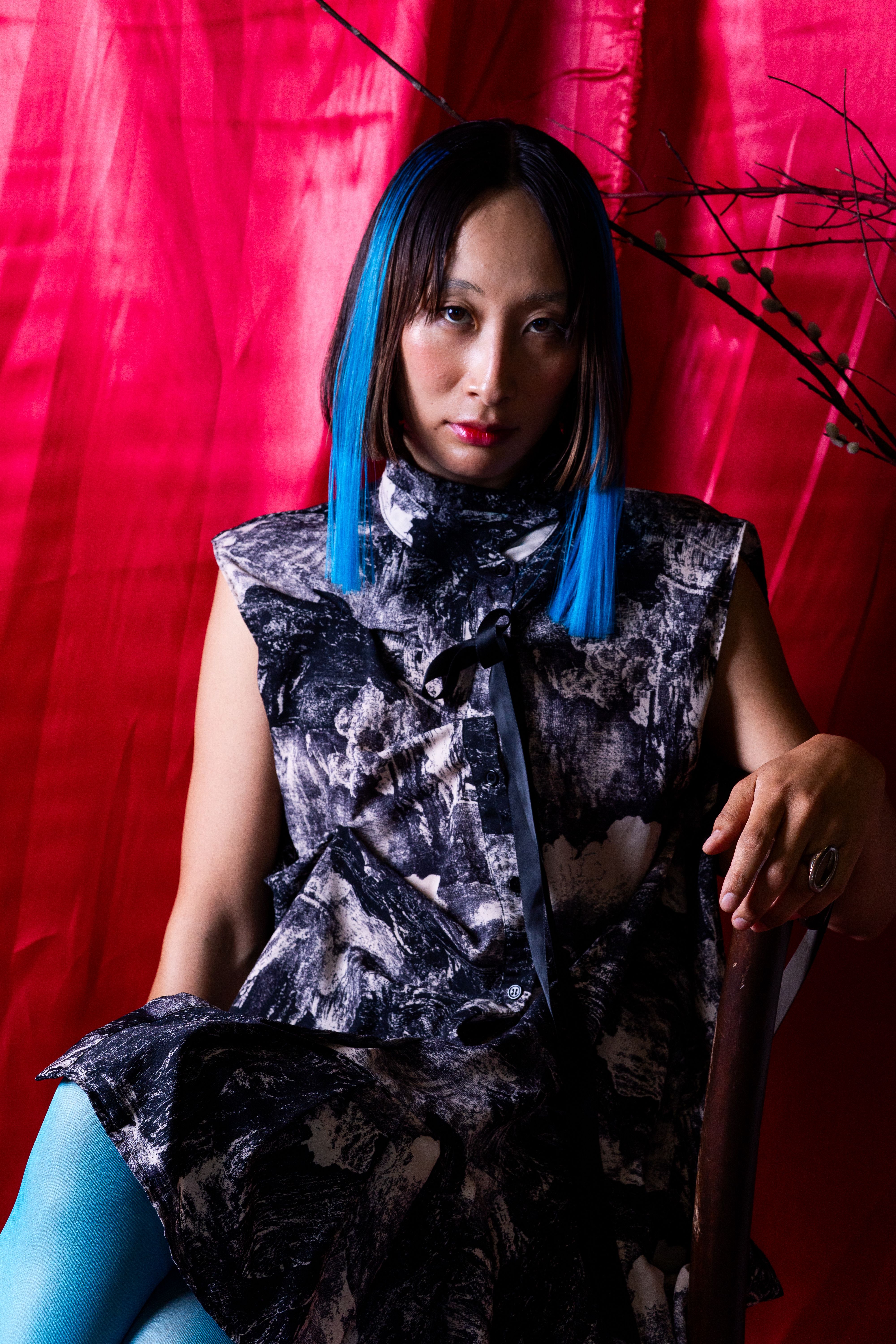 Stylish Asian model with blue hair posing against a vibrant red backdrop, showcasing bold fashion.