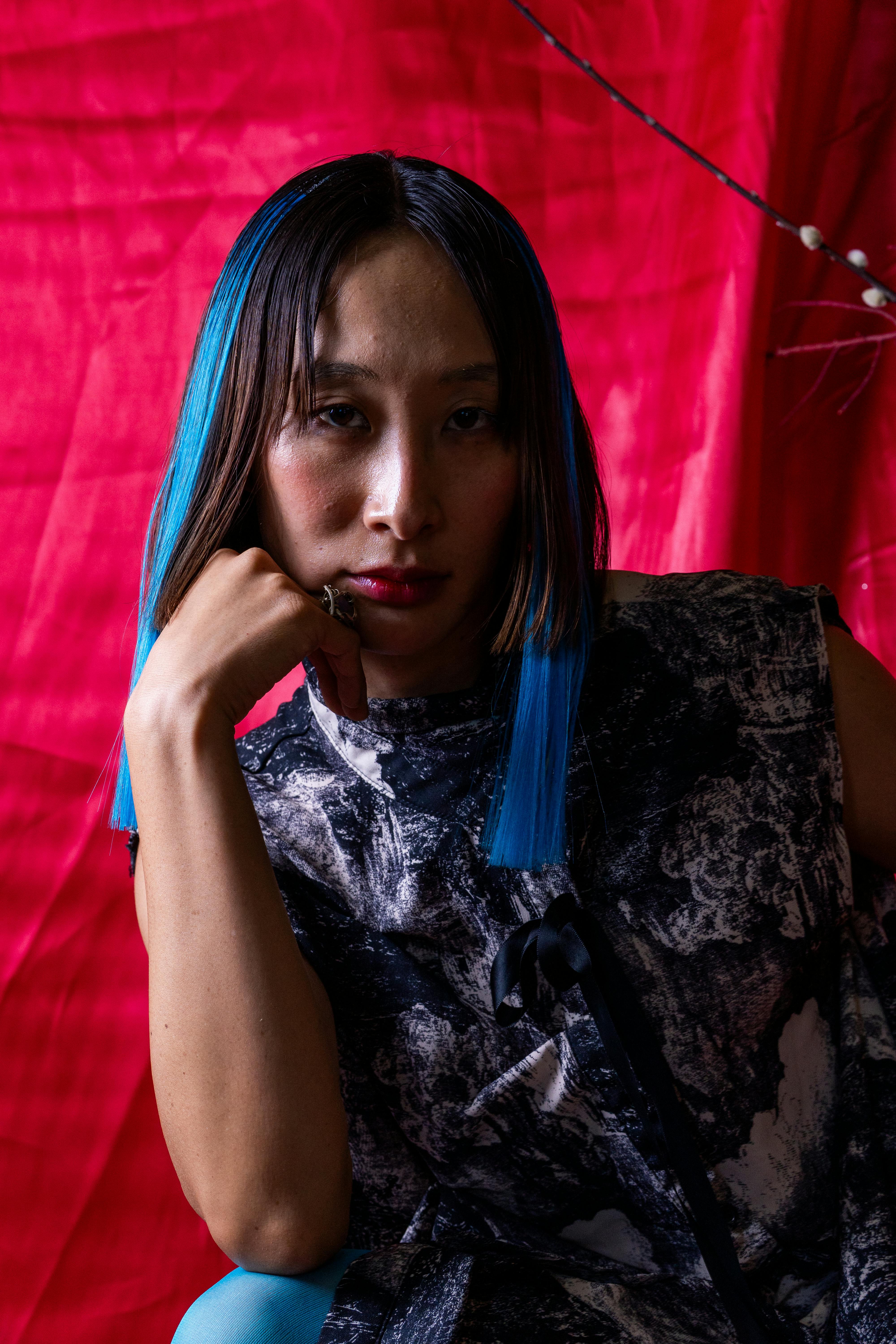 Striking portrait of woman with blue hair in artistic pose against red backdrop.