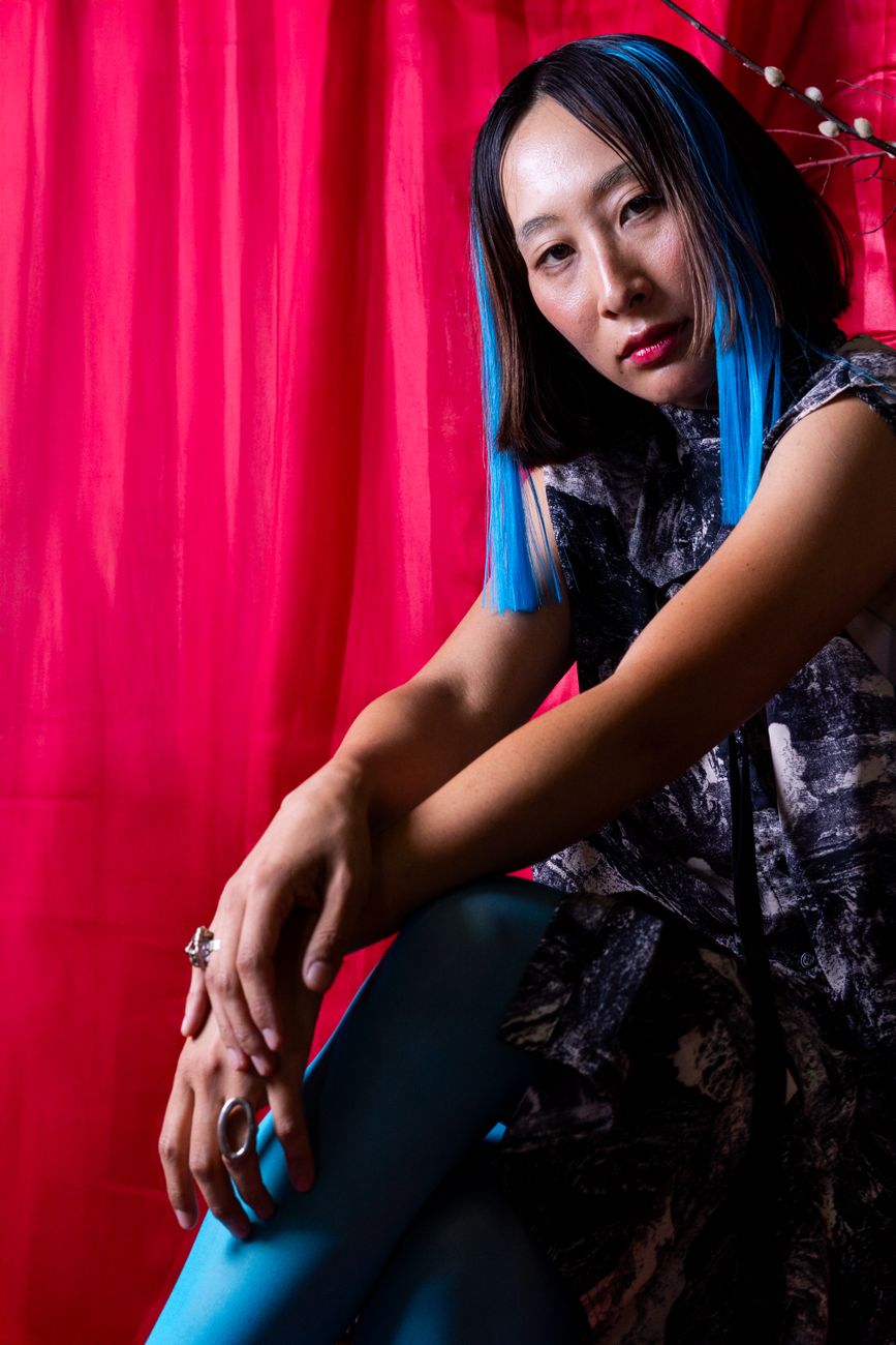 Fashionable woman with blue hair in a striking pose against a red backdrop.