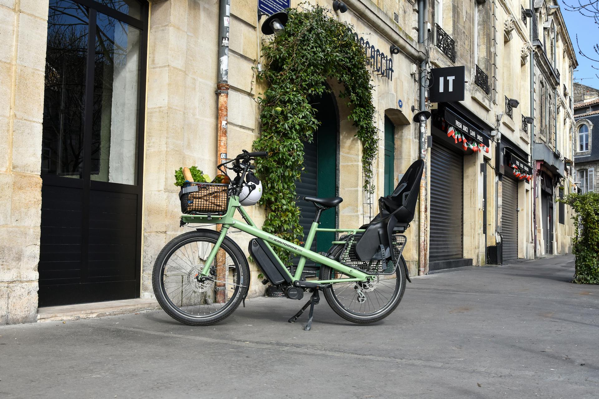 A green cargo e-bike with groceries parked outside a French building on a sunny day.