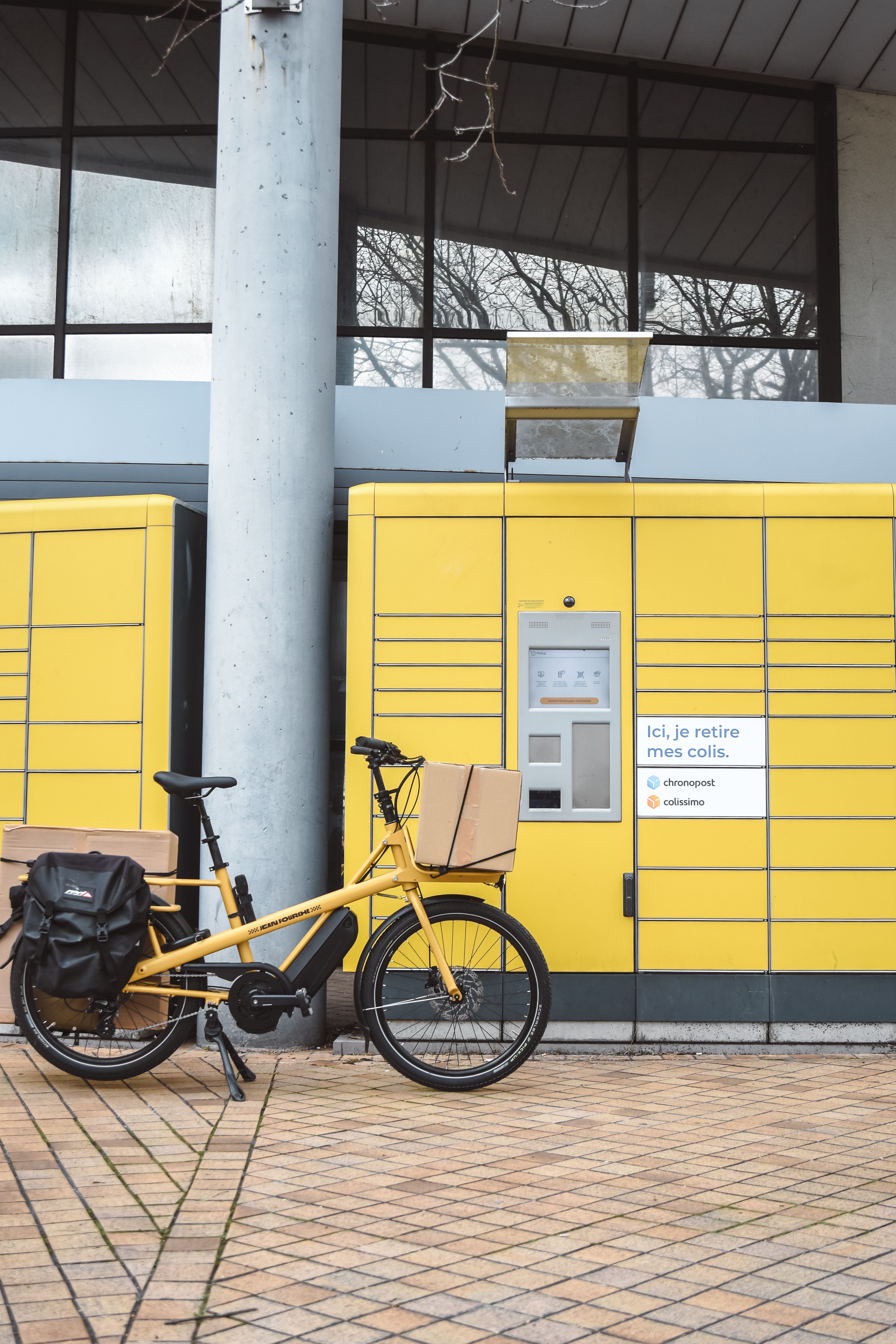 Yellow cargo bike with parcels parked by a parcel locker station in France.
