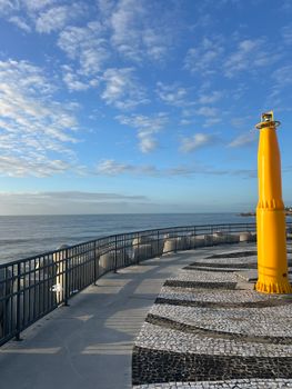 Scenic coastal promenade featuring a yellow beacon overlooking the ocean under a blue sky.