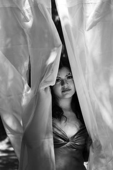 Artistic black and white portrait of a woman peeking through sheer drapes, conveying mystery.