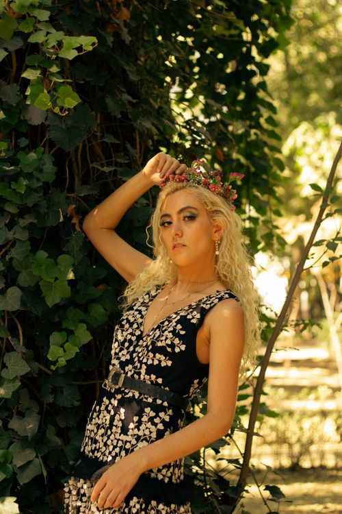 Free Blonde woman in black floral dress poses gracefully outdoors in a sunlit garden. Stock Photo