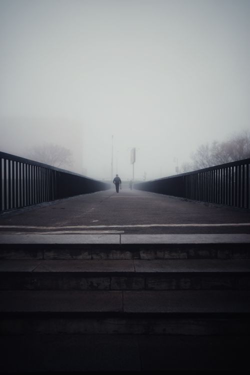 Free Silhouette of a person walking alone on a foggy footbridge creating a moody atmosphere. Stock Photo