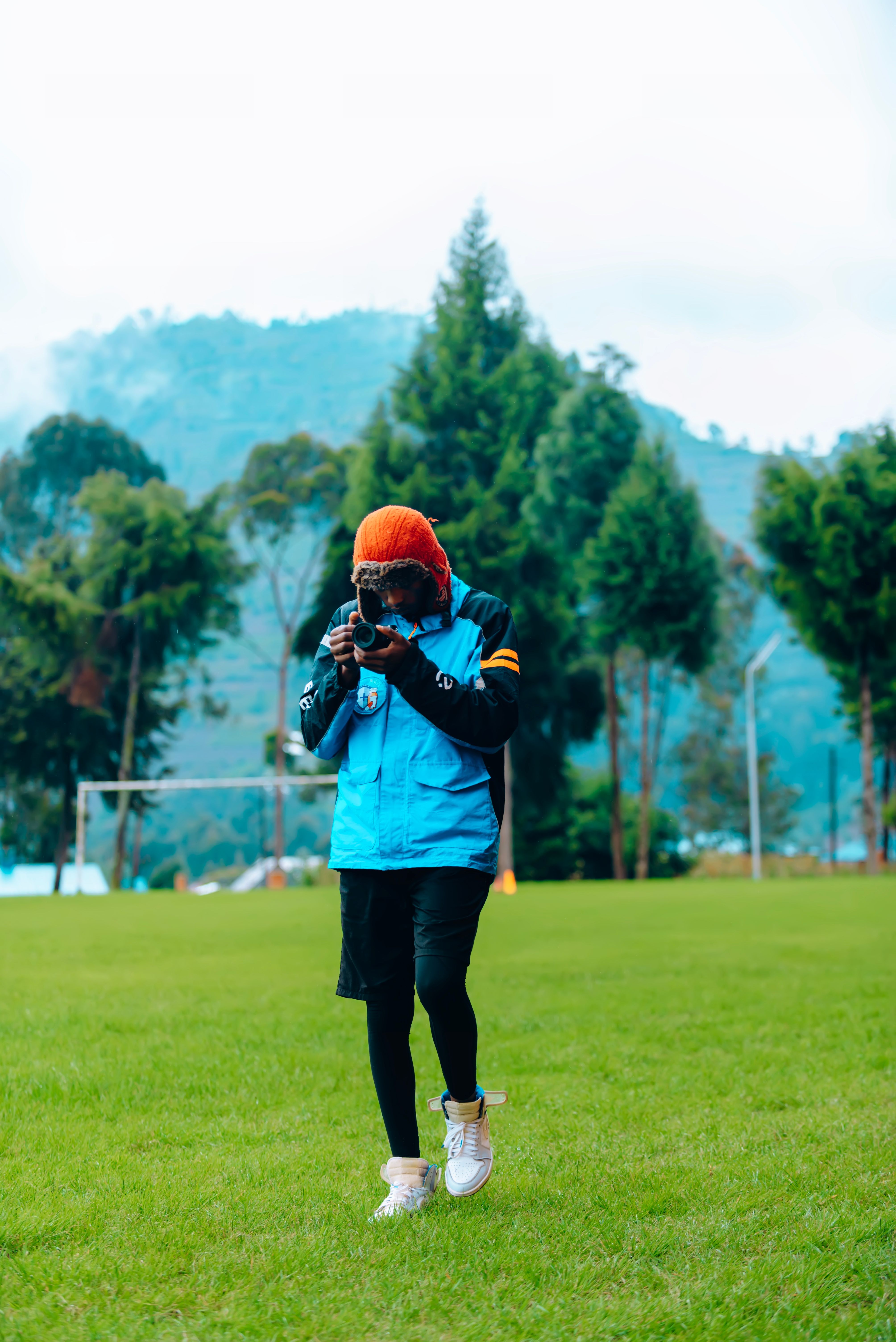 A young photographer in vibrant outerwear captures nature on a field, surrounded by mountains.