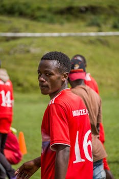 Teen soccer player in red jersey on field, looking back towards camera, teammates in background.