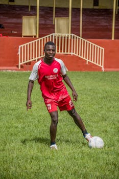 Young soccer player in red uniform dribbling a ball on a grass field during the day.