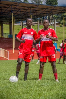 Two young African soccer players in red uniforms on a grassy field, ready for practice.