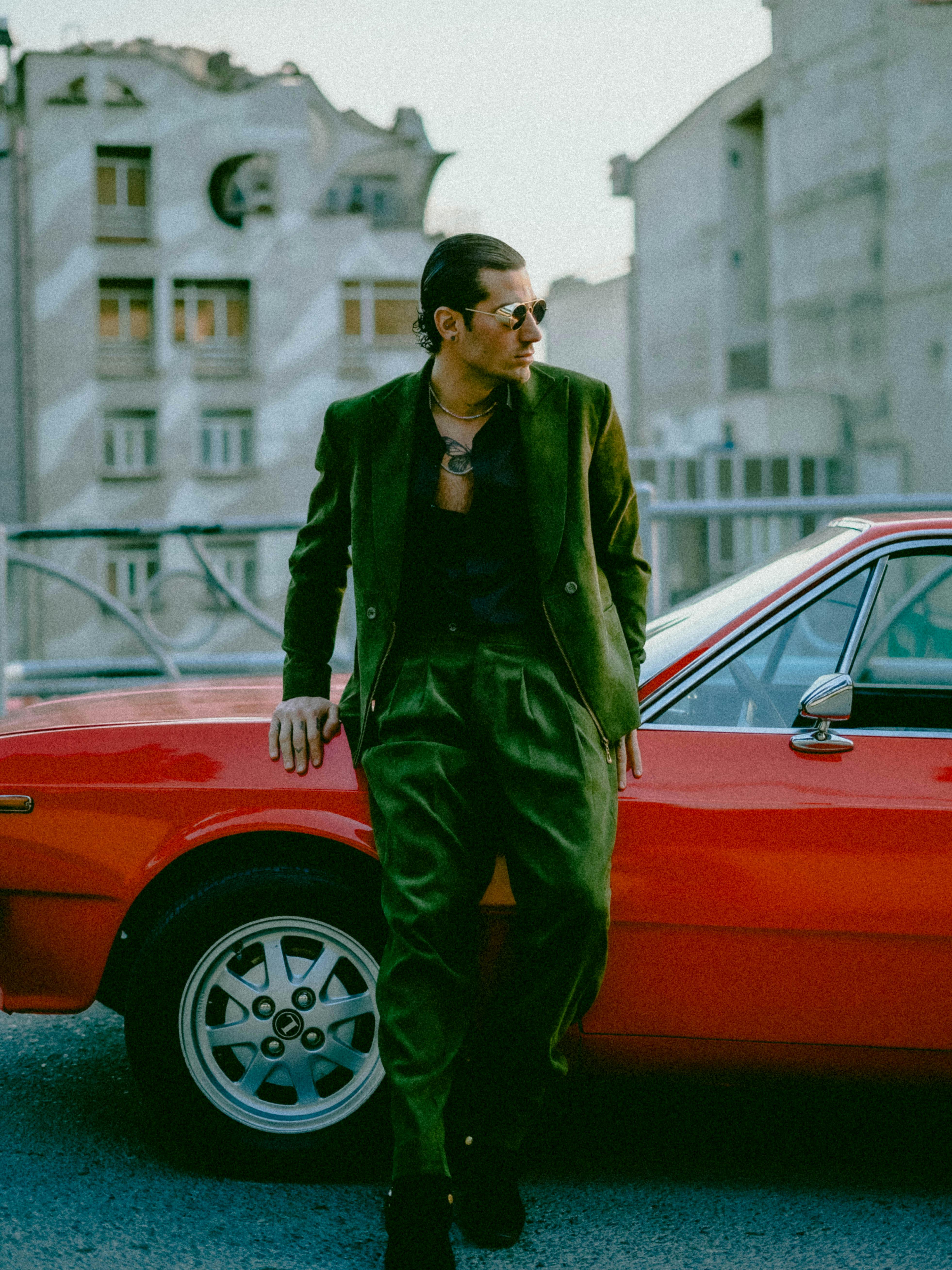 Fashionable man in green suit leaning on a classic red car in an urban setting.