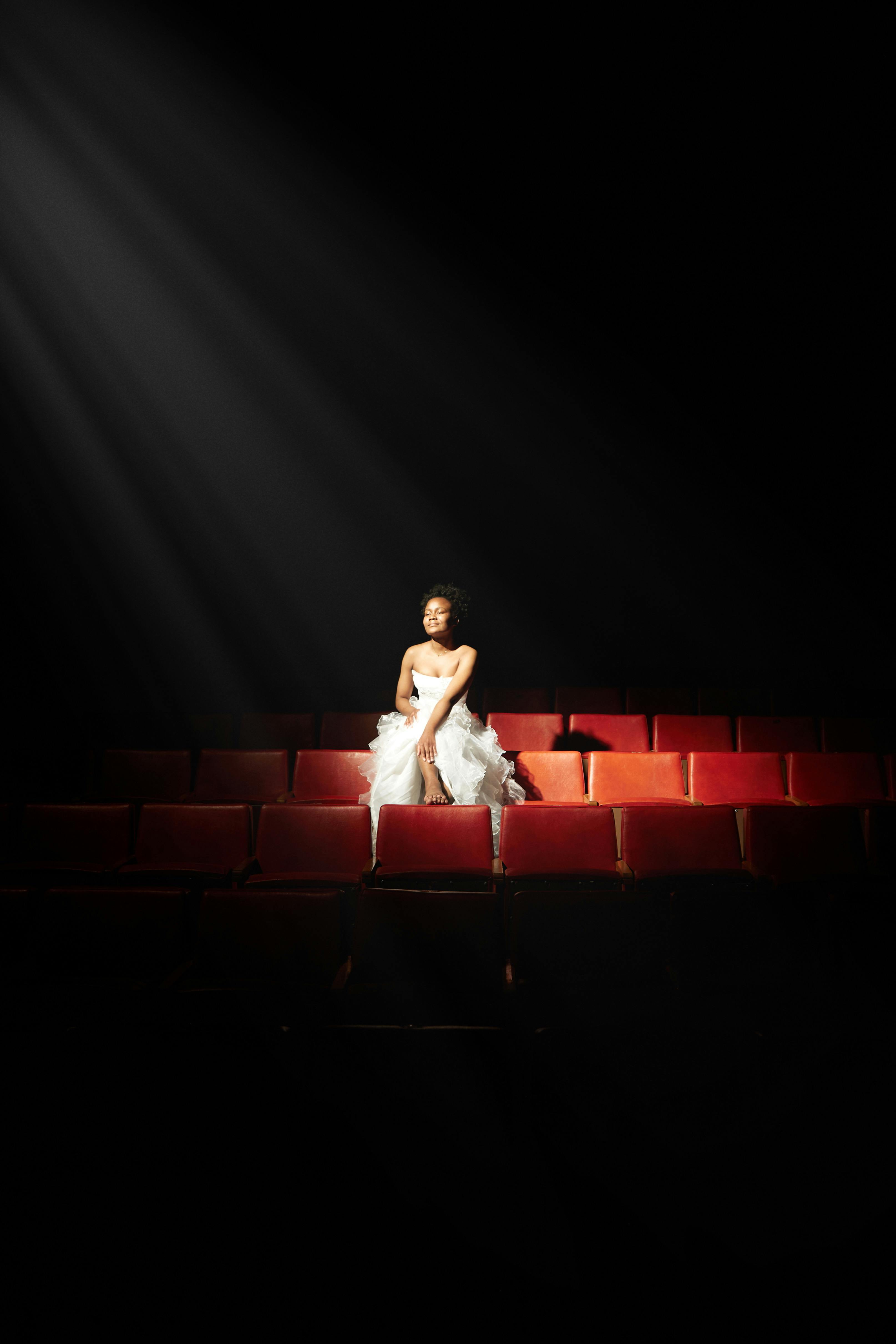 Free Woman in white dress sits in spotlight on red theater seats. Stock Photo