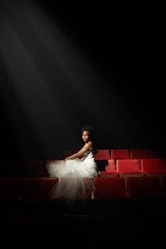 Elegant woman in white gown seated in theater, illuminated by spotlight. Dramatic scene.