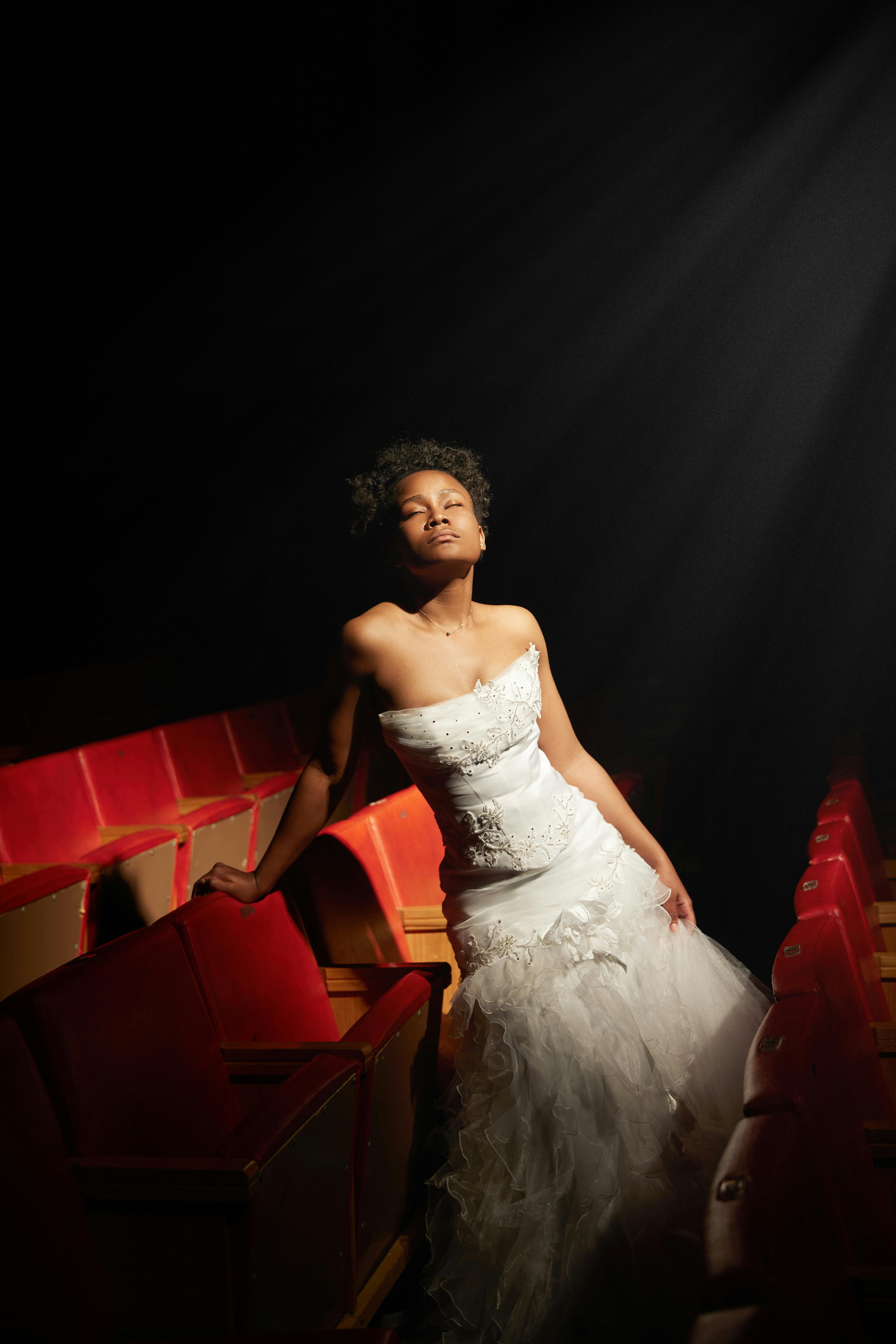 Ücretsiz Gelinlik giymiş bir kadın, spot ışıklarının aydınlattığı bir tiyatroda hayalperest bir şekilde poz veriyor. Stok Fotoğraflar