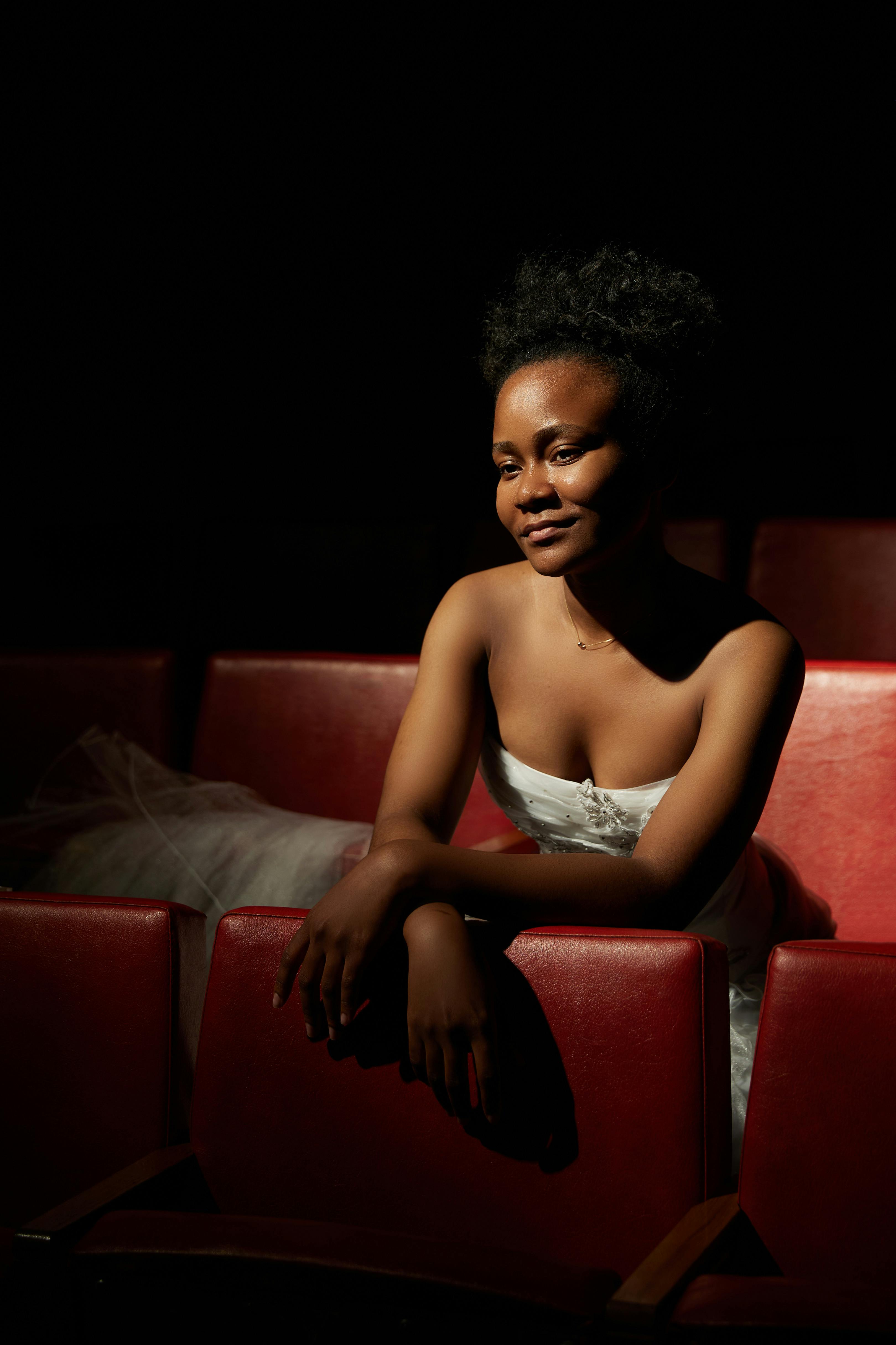 Free Graceful woman sits in dimly lit theater wearing a white gown, exuding elegance. Stock Photo