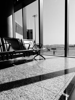 A moody black and white photo of empty airport seating against large windows with sunlit reflections.