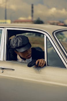 A woman with a black cap leans out of a vintage car window holding a camera.