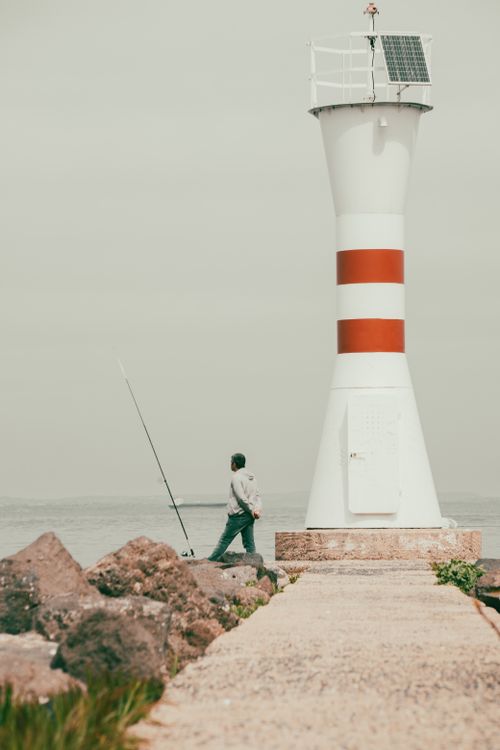 Free A lone fisherman with a rod beside a red and white striped lighthouse on a rocky shore. Stock Photo