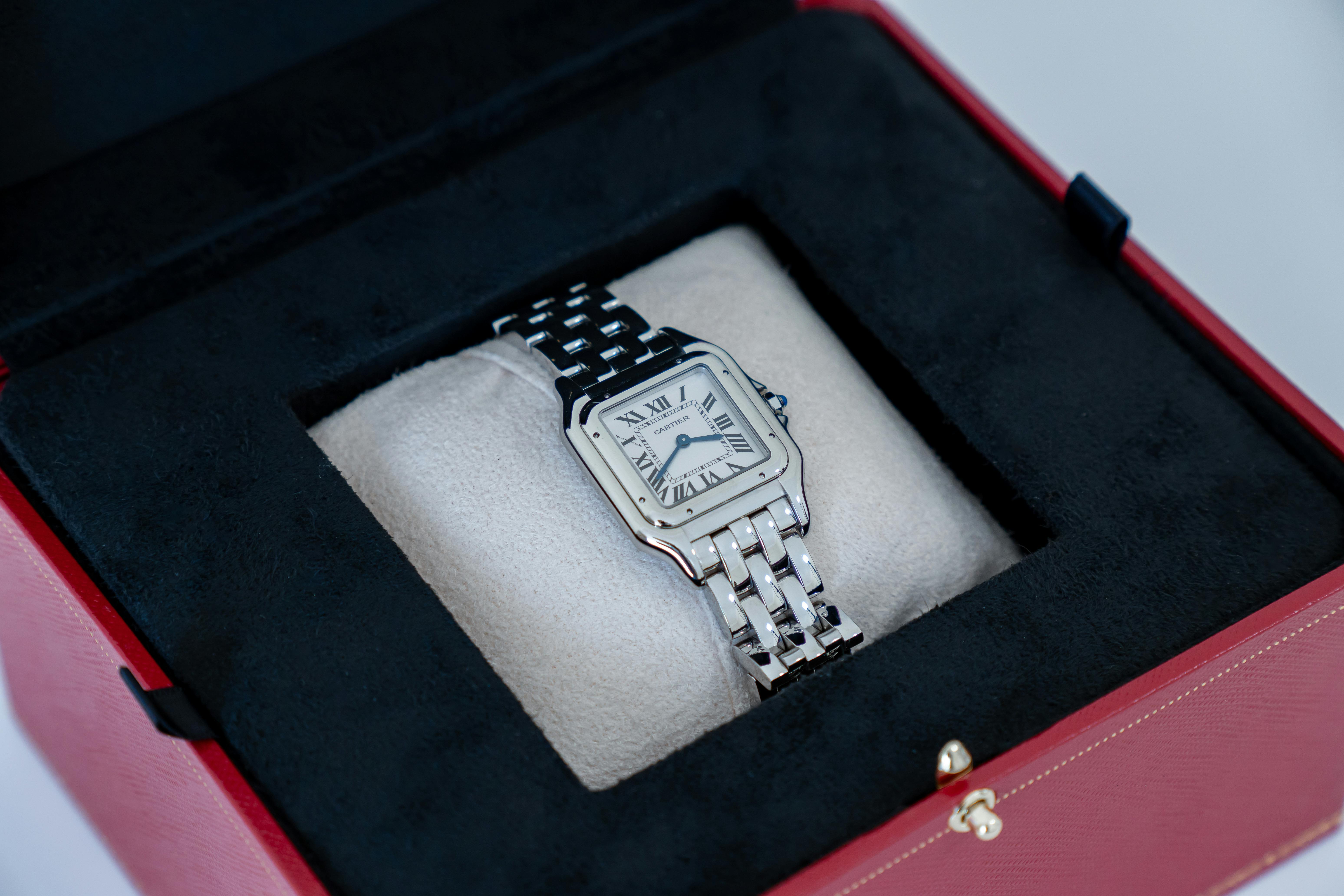 A silver Cartier Panthère watch with a metal link bracelet sits on a beige cushion inside an open red jewelry box.