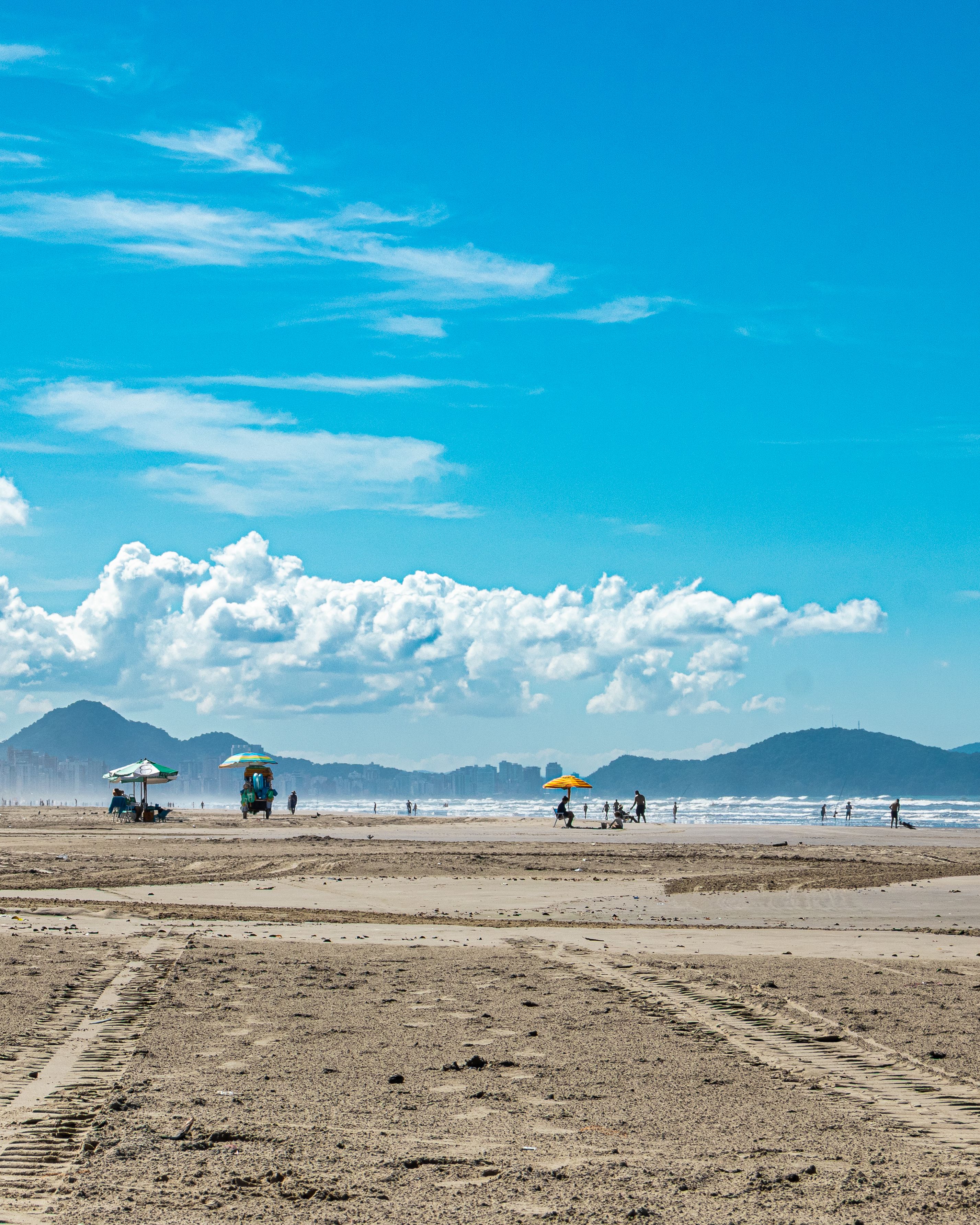Sunny day at Praia Grande with beachgoers, blue sky, and distant mountains.