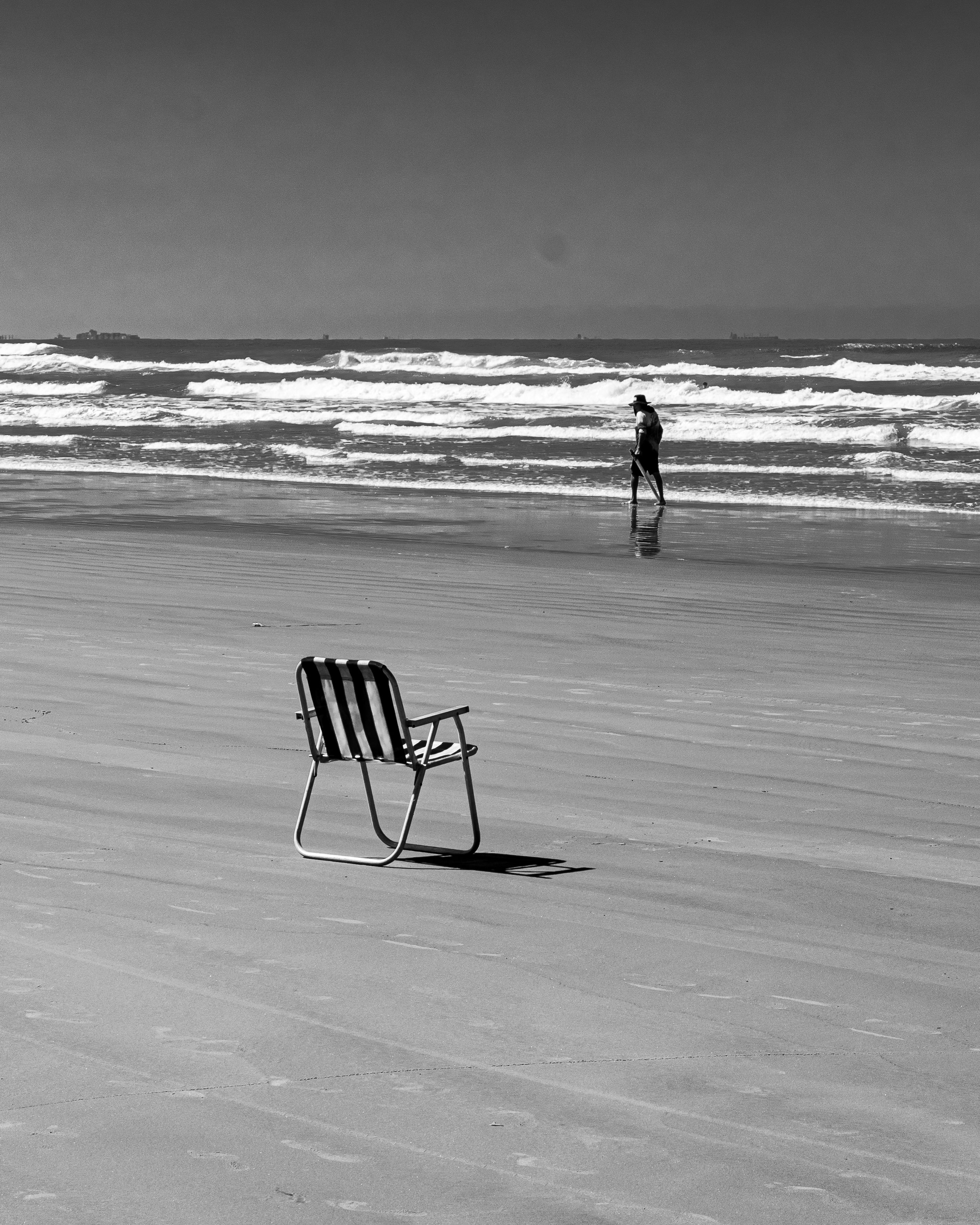 Black and white photo of a beach chair and a lone walker by the ocean at Praia Grande, Brazil.
