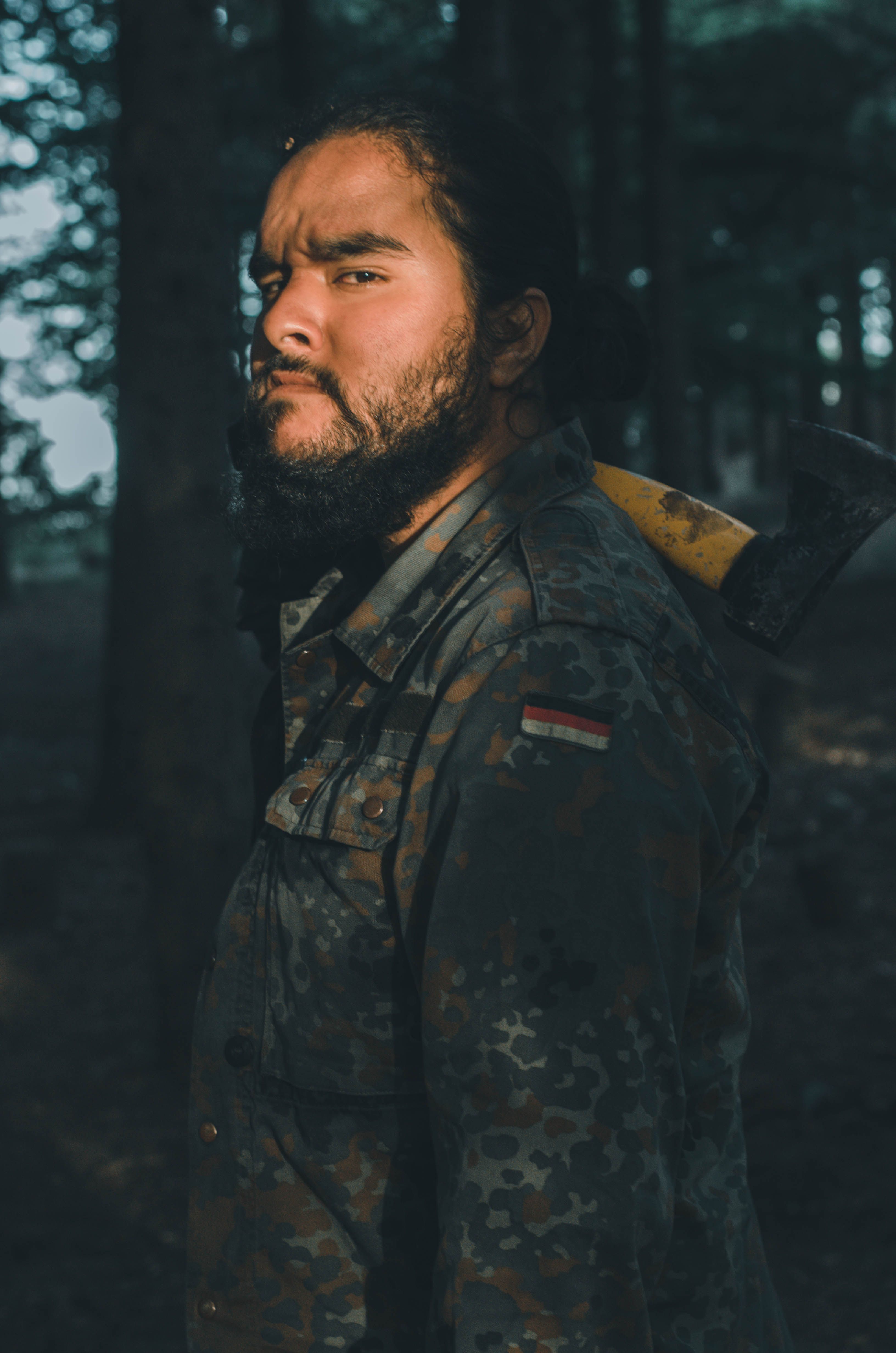 A bearded man in a camouflage jacket stands in a sunlit forest, exuding a rugged outdoor vibe.