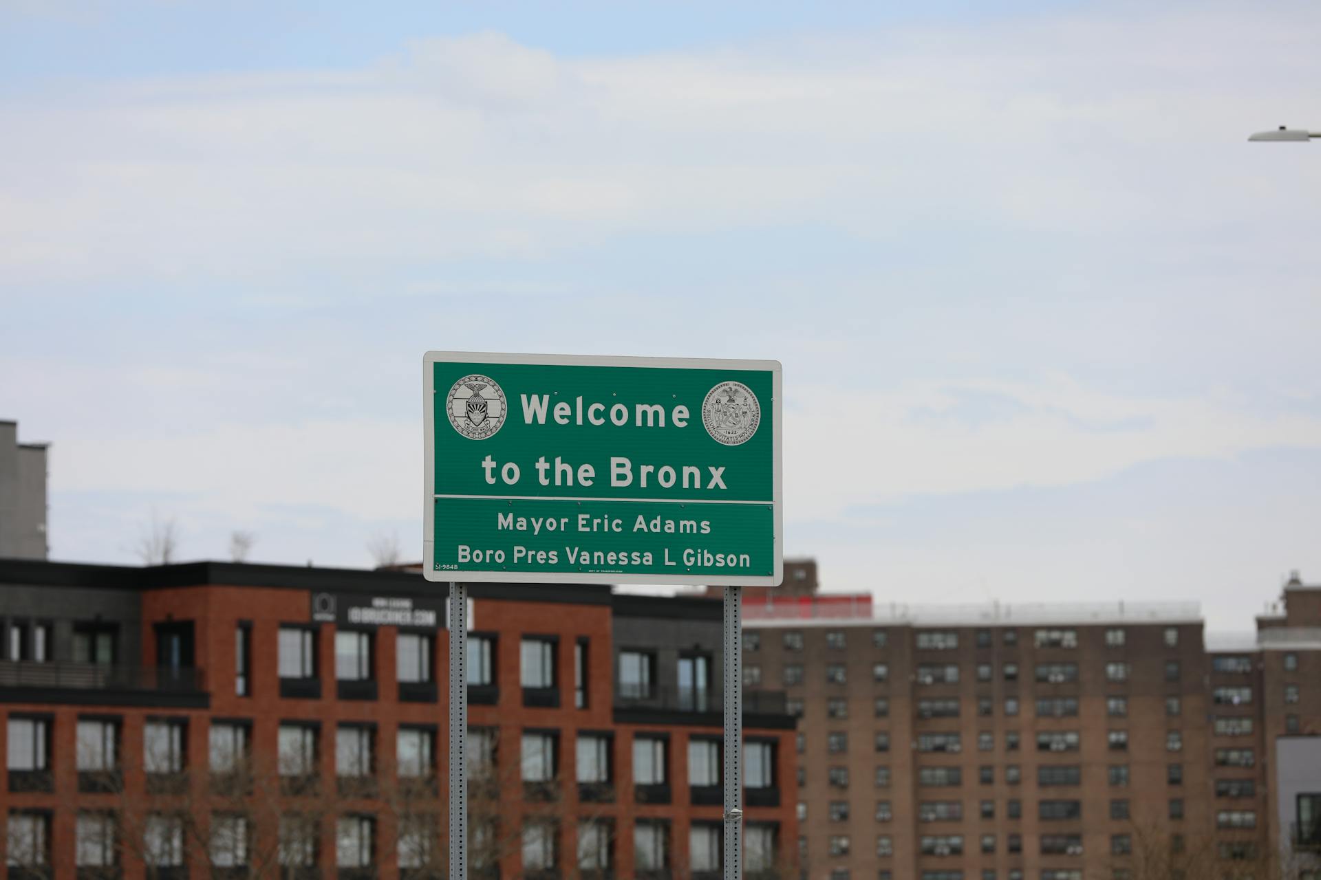 A cityscape featuring the Welcome to the Bronx sign in New York City, with residential buildings in the background.