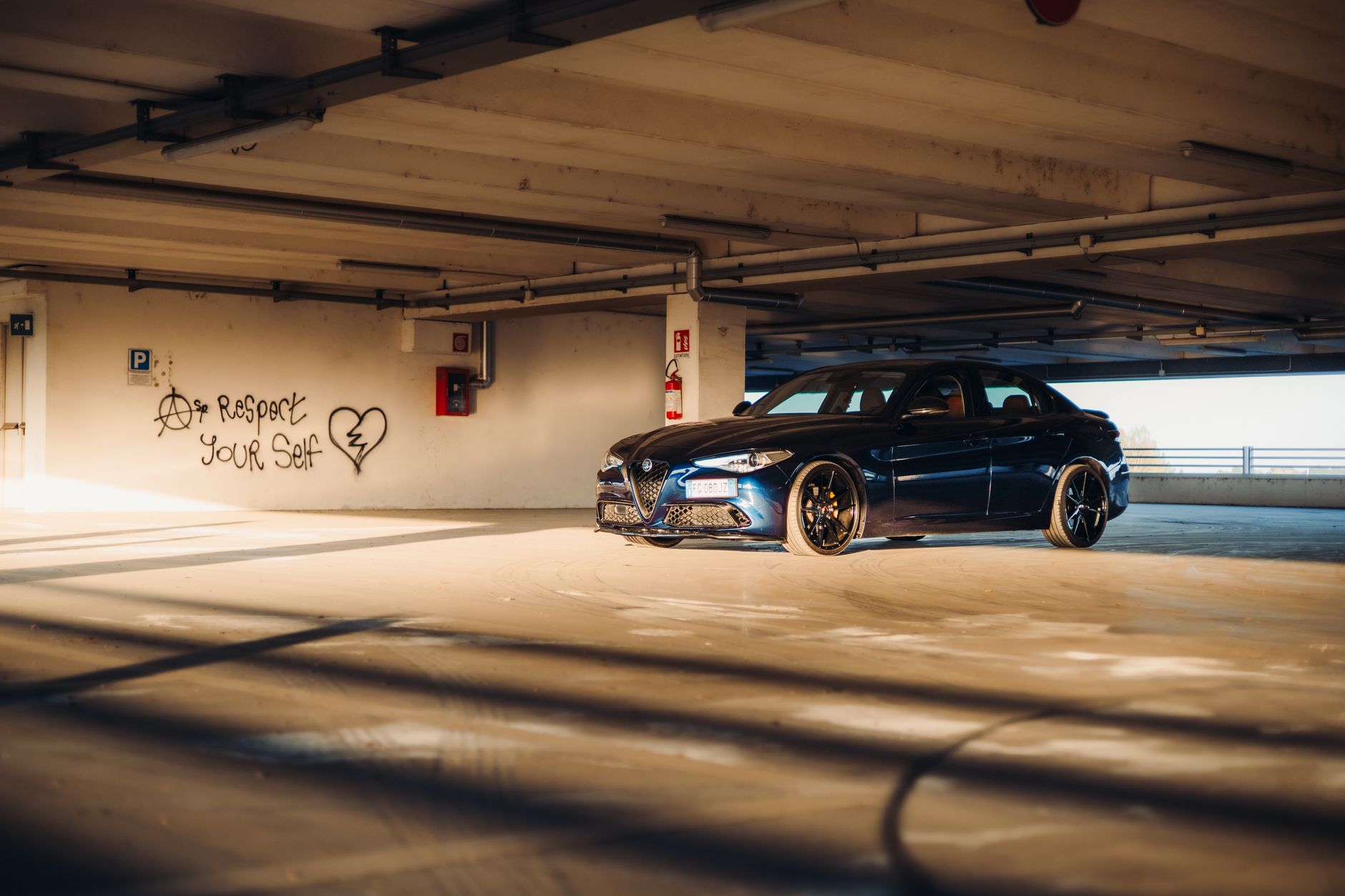 Luxury car parked in sunlit urban garage with graffiti art on walls.