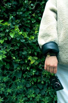 Close-up of person wearing beige coat, holding a camera against a leafy green background.